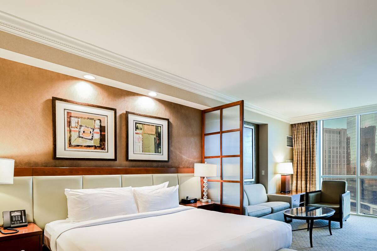 A hotel room with a large bed, a sofa, a desk, and a view of the city through large windows.