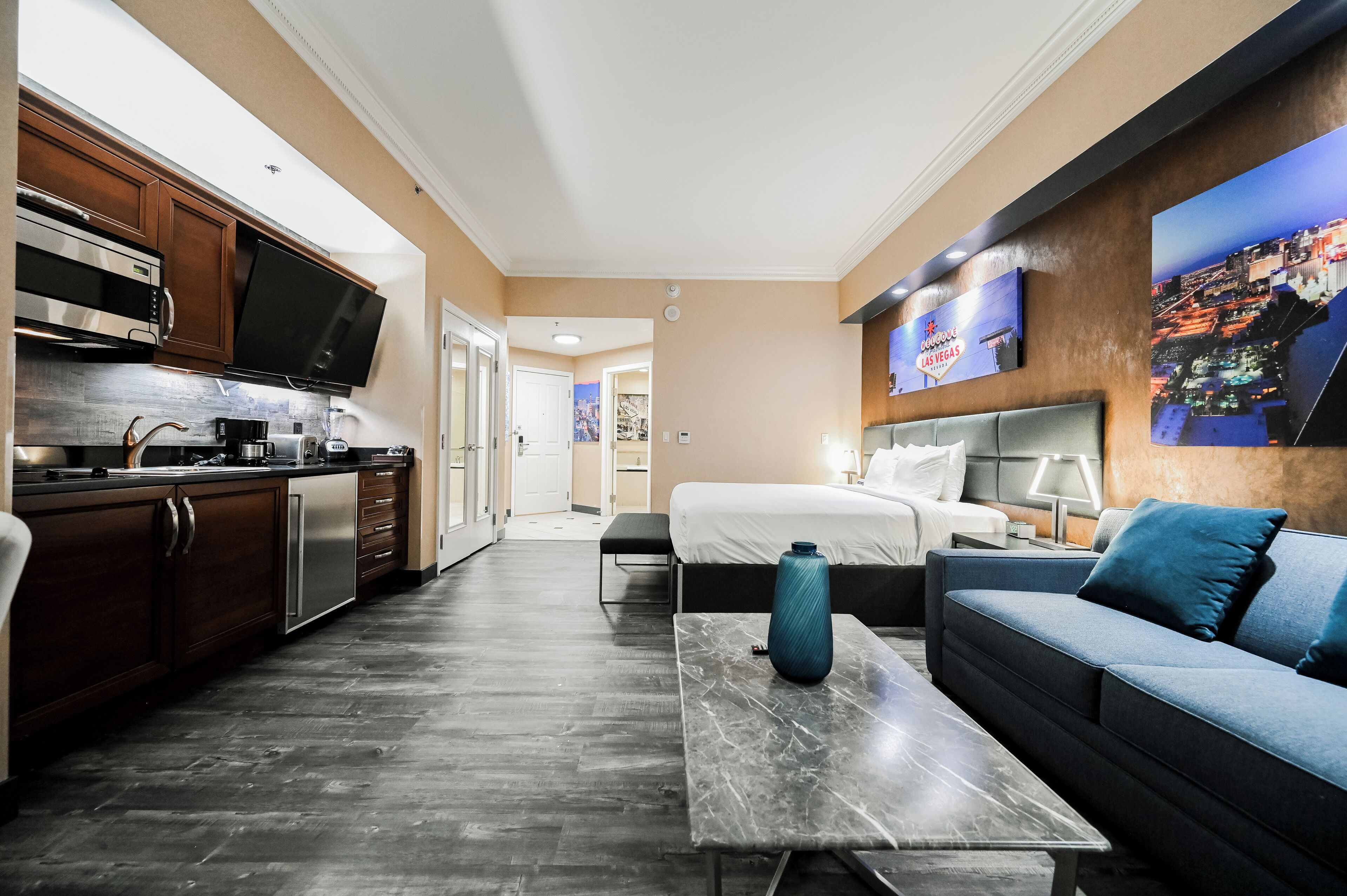 Ein modernes Hotelzimmer mit Küchenzeile, einem Bett mit Kopfteil, einer Couch, einem Couchtisch und einem an der Wand montierten Fernseher.