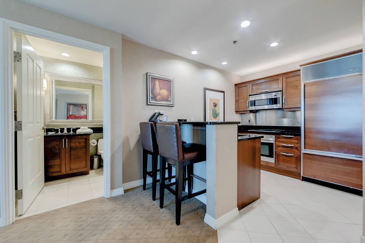 A modern kitchen with wooden cabinets, a built-in microwave, and a refrigerator. There is a small island with bar stools and a painting on the wall.