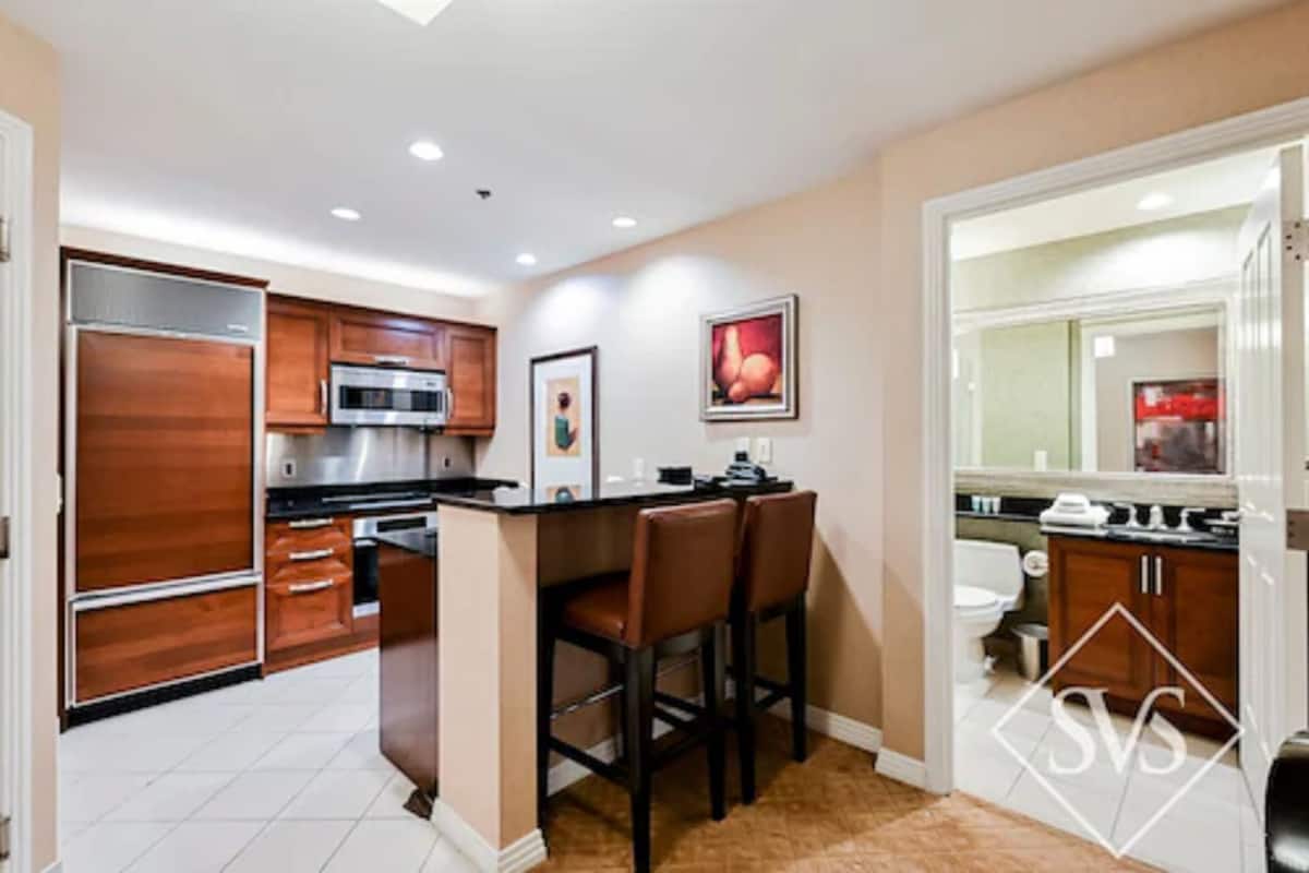 A modern kitchen with wooden cabinets, a stainless steel refrigerator, and a bar area with two stools.