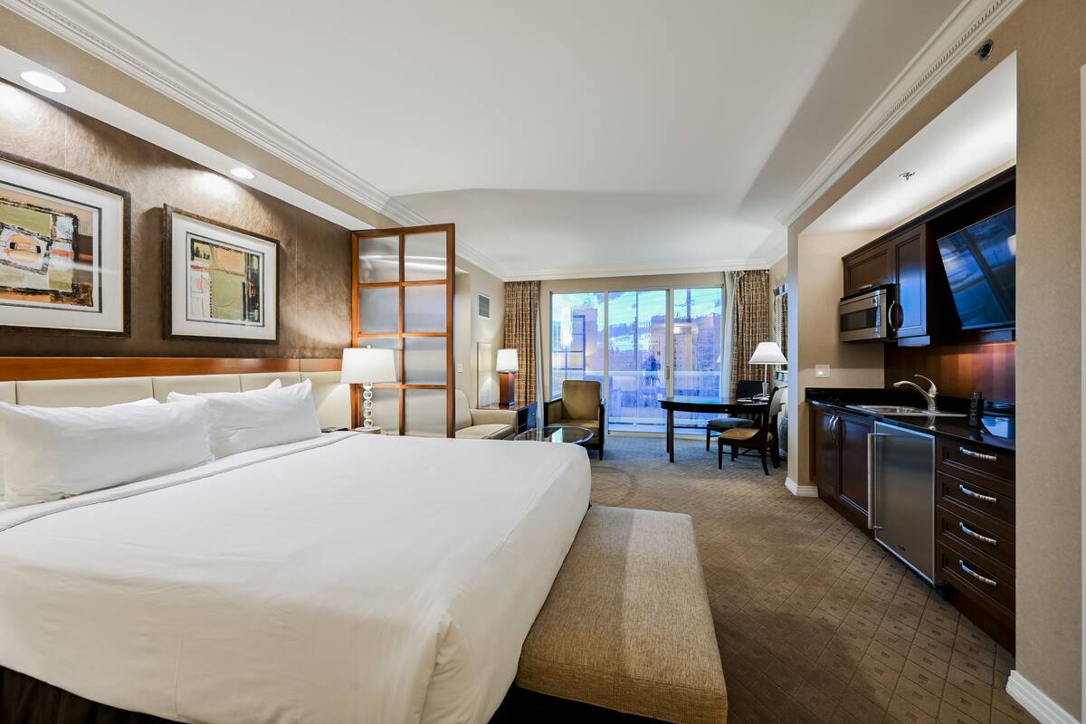 A hotel room with a large bed, a desk, a chair, a television, and a kitchenette.