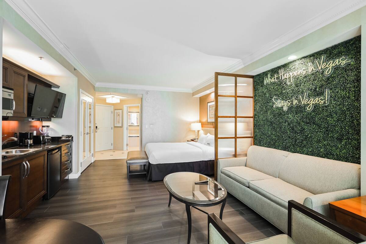 A hotel room with a kitchenette, a bed, a sofa, and a green wall with a neon sign.