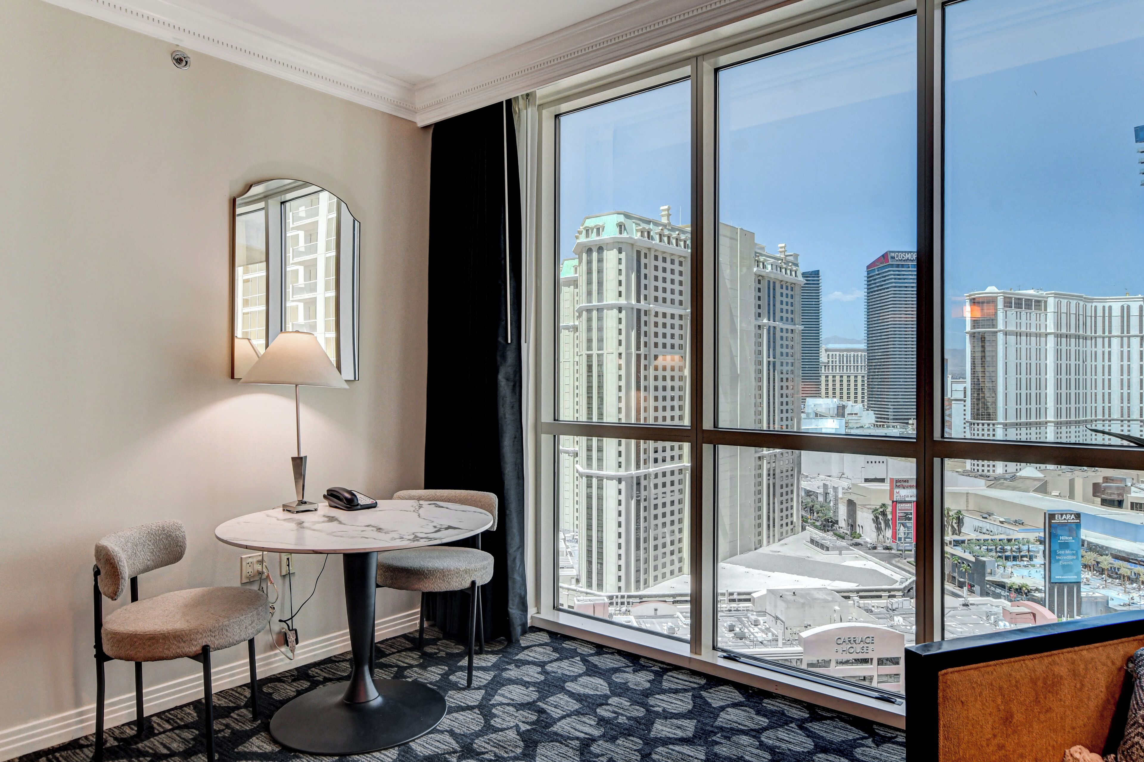 Hotelkamer met een groot raam met uitzicht op de stad, een ronde tafel met twee stoelen, een lamp en een spiegel aan de muur.