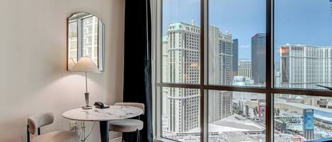Hotelkamer met een groot raam met uitzicht op de stad, een ronde tafel met twee stoelen, een lamp en een spiegel aan de muur.