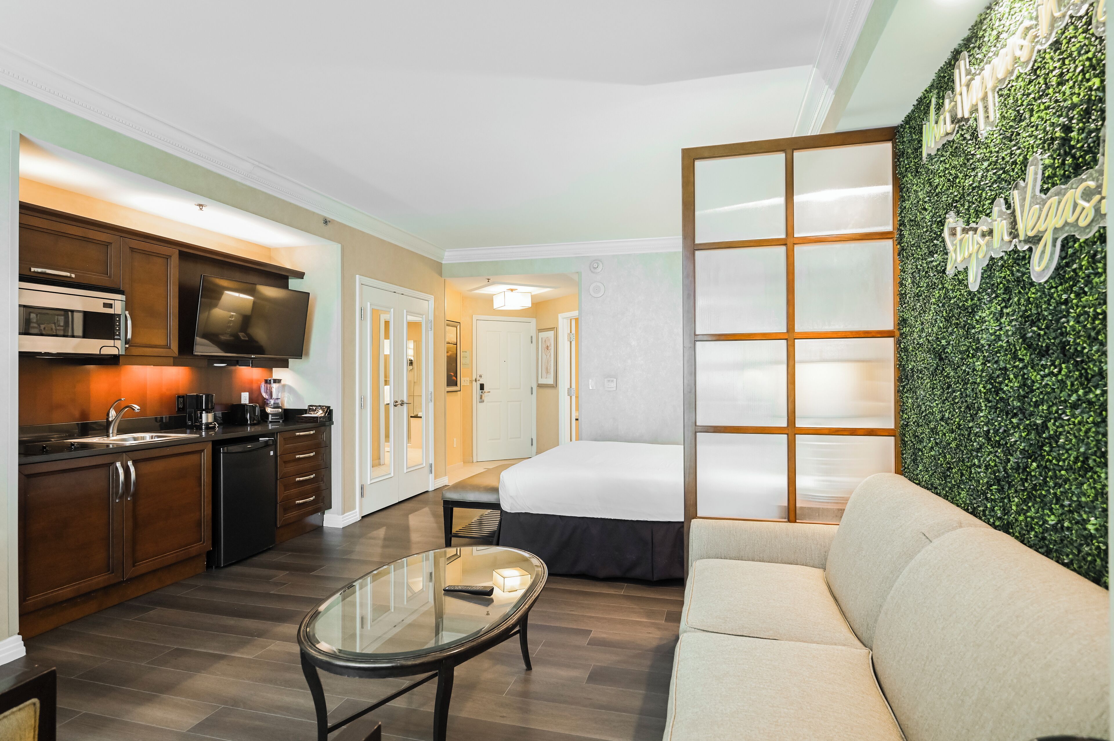 A compact hotel room with a kitchenette, a bed, a sofa, and a decorative wall with greenery.