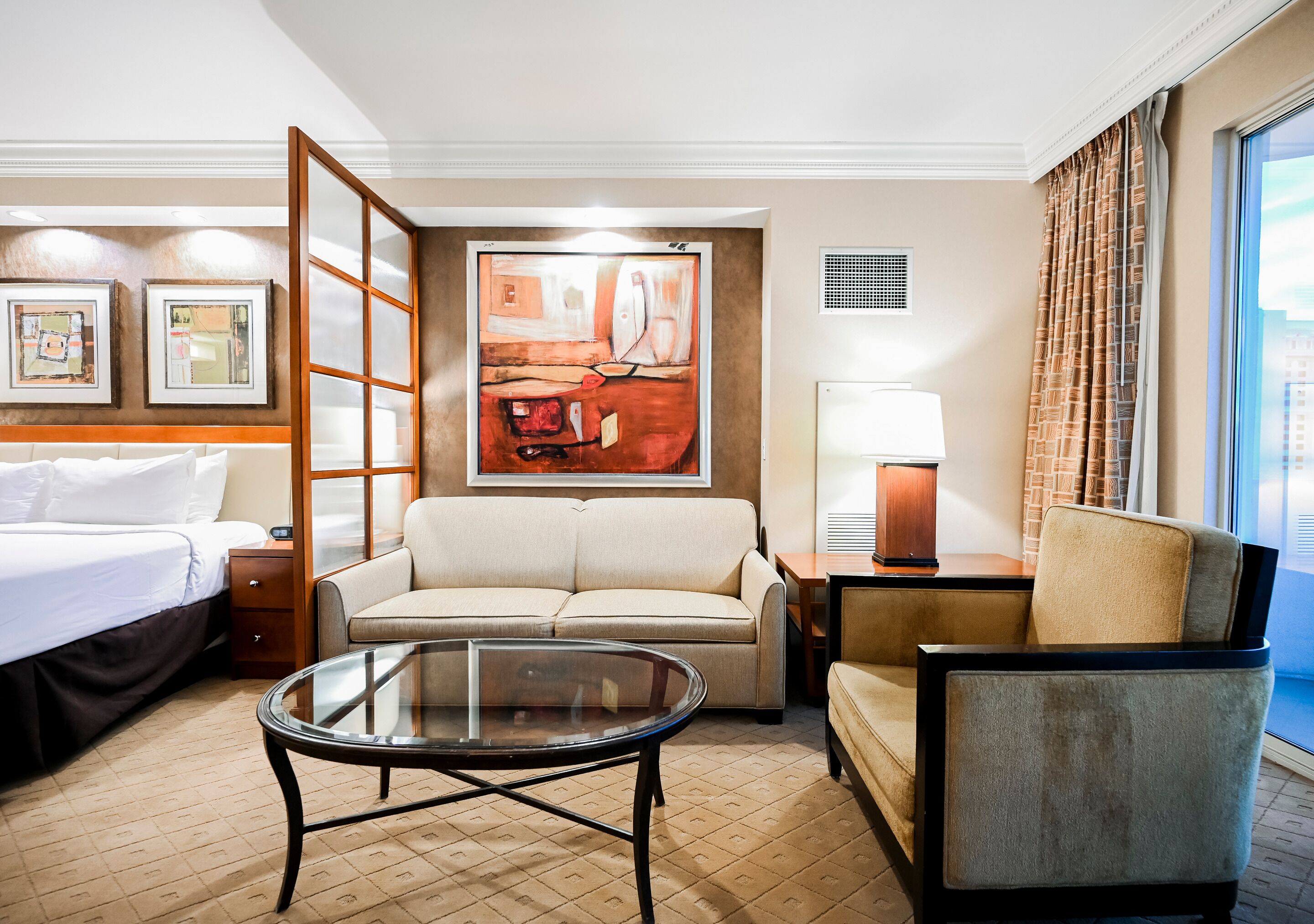 A hotel room with a bed, a sofa, a chair, a glass coffee table, a painting on the wall, and a window with curtains.