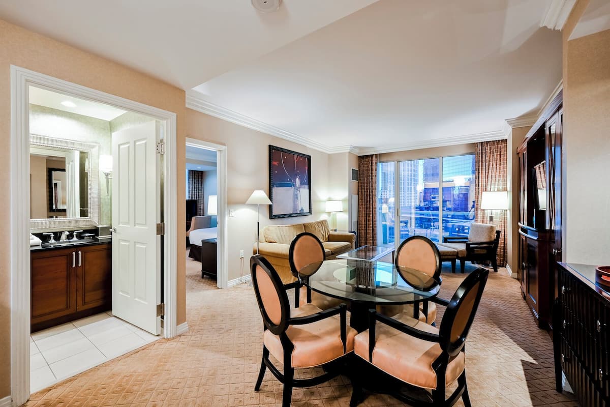 A hotel room with a dining area featuring a round glass table and leather chairs, a large window with a city view, and a separate bedroom visible through an open door.