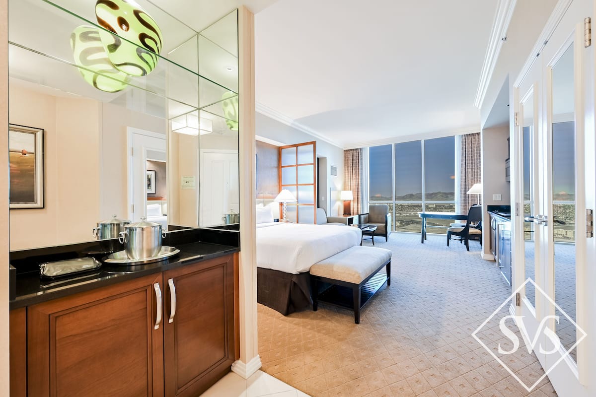 A hotel room with a large bed, a desk, a chair, a balcony with a view, and a kitchen area with a stove and pots.