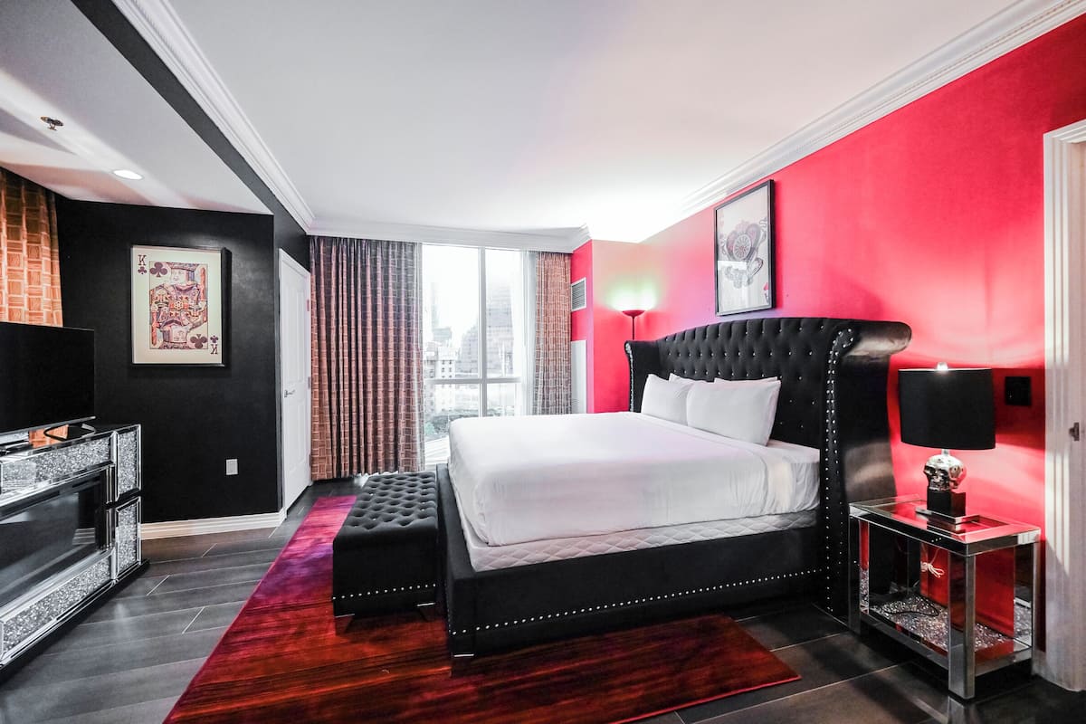A modern bedroom with a black and white bed, a red accent wall, and a city view through large windows.