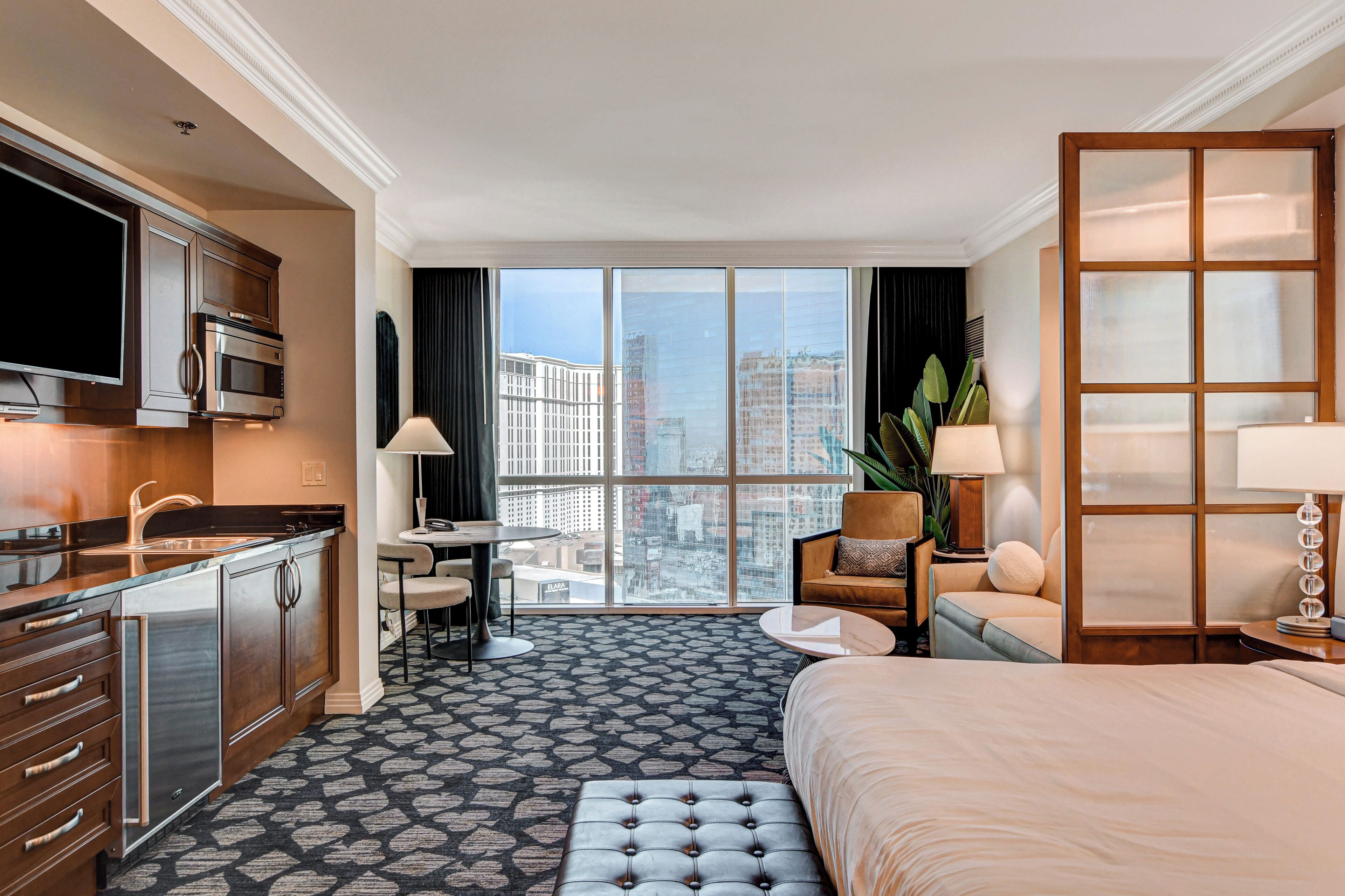Una habitación de hotel con cama, escritorio, silla, sofá, televisión y vista a la ciudad a través de amplios ventanales.