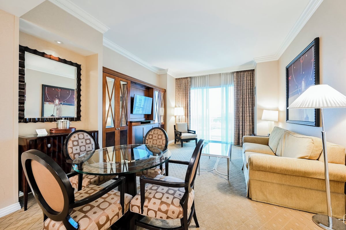 A hotel room with a glass dining table, four chairs, a sofa, a TV, and a large window with curtains.