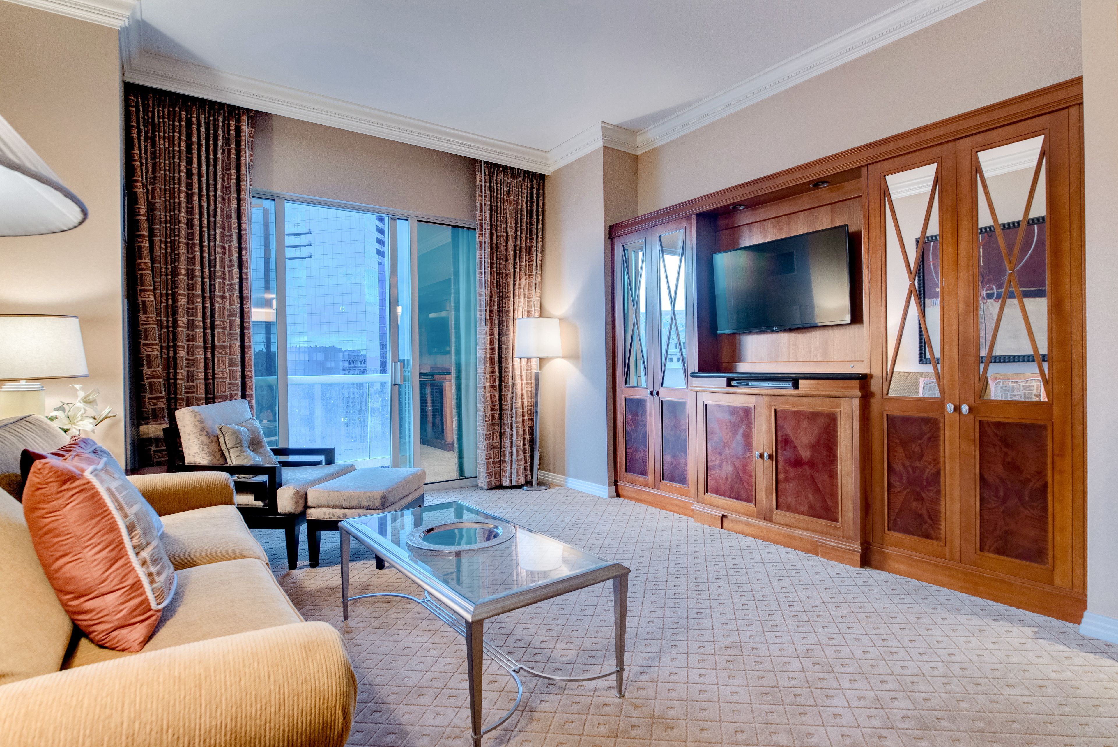 Une chambre d’hôtel équipée d’un canapé, d’un fauteuil, d’une table basse en verre, d’une télévision et d’une armoire en bois.