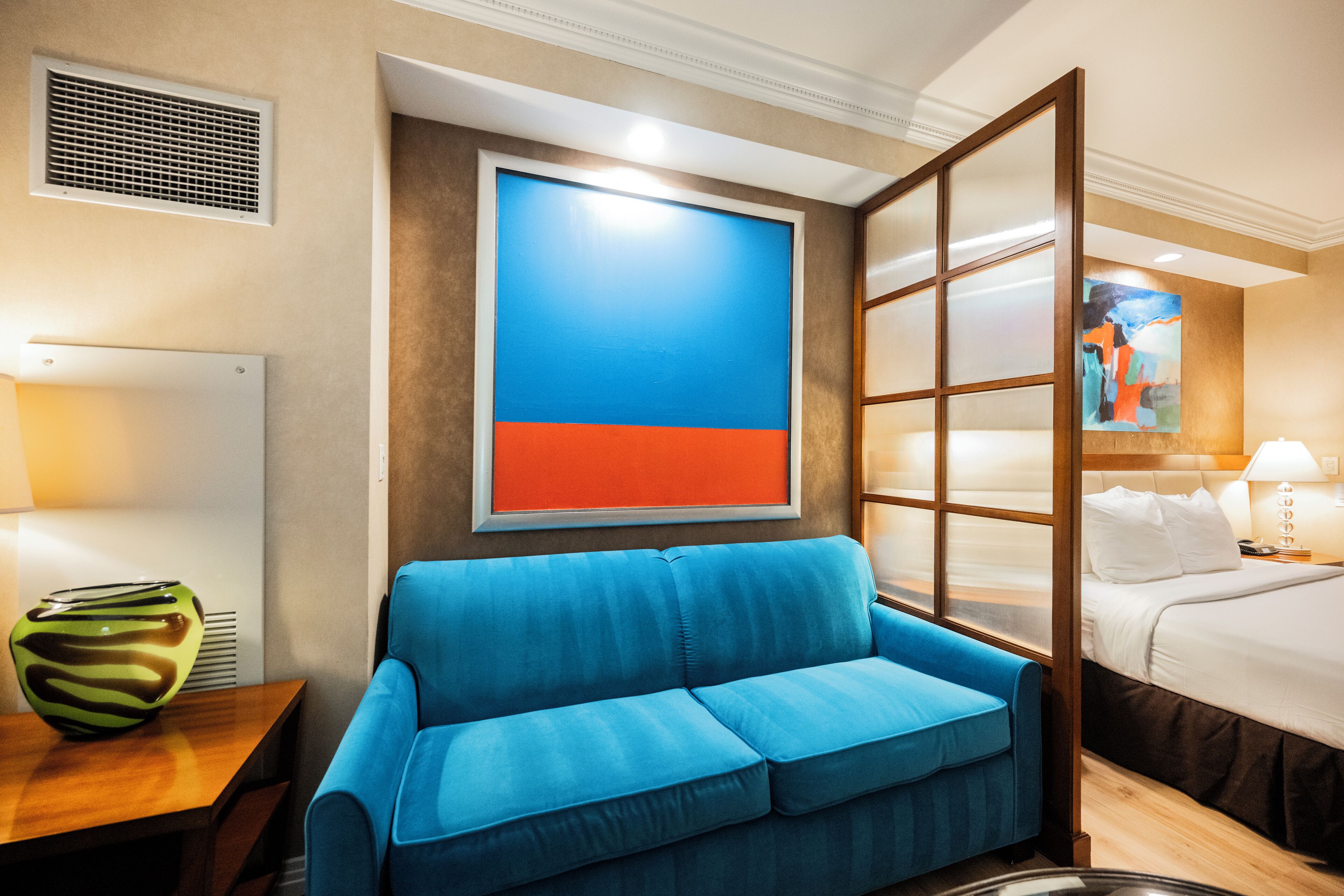 Habitación de hotel con un sofá azul, una cama, un cuadro y una rejilla en la pared.