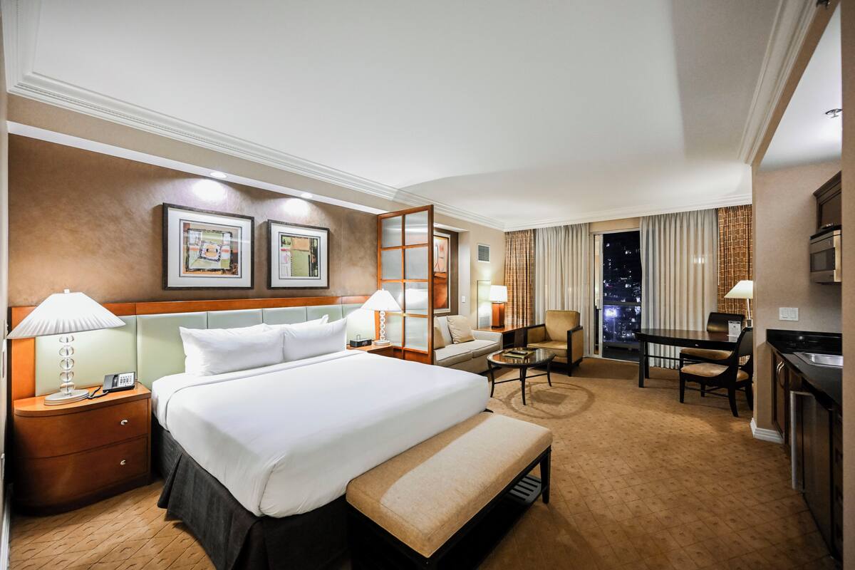 A hotel room with a large bed, bedside tables, a desk, a chair, and a view of the city at night.