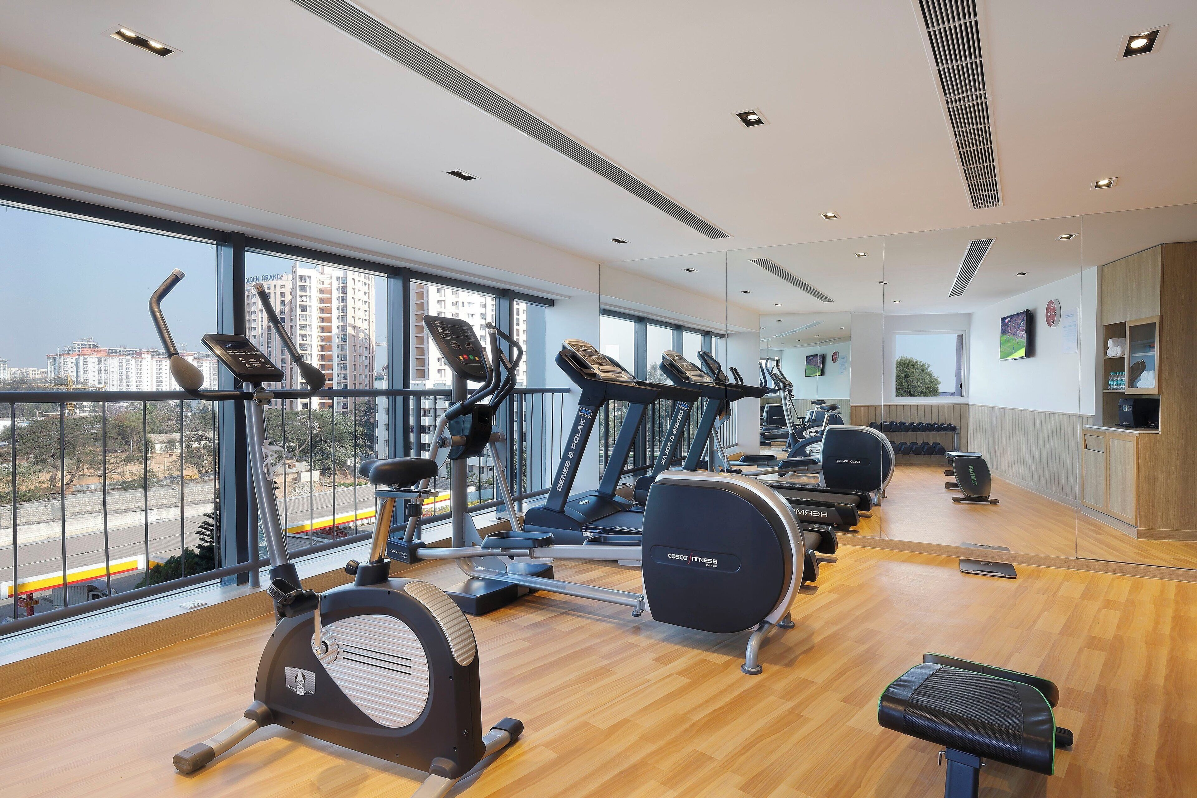 A gym with cardio machines, a mirrored wall, and a view of buildings through large windows.