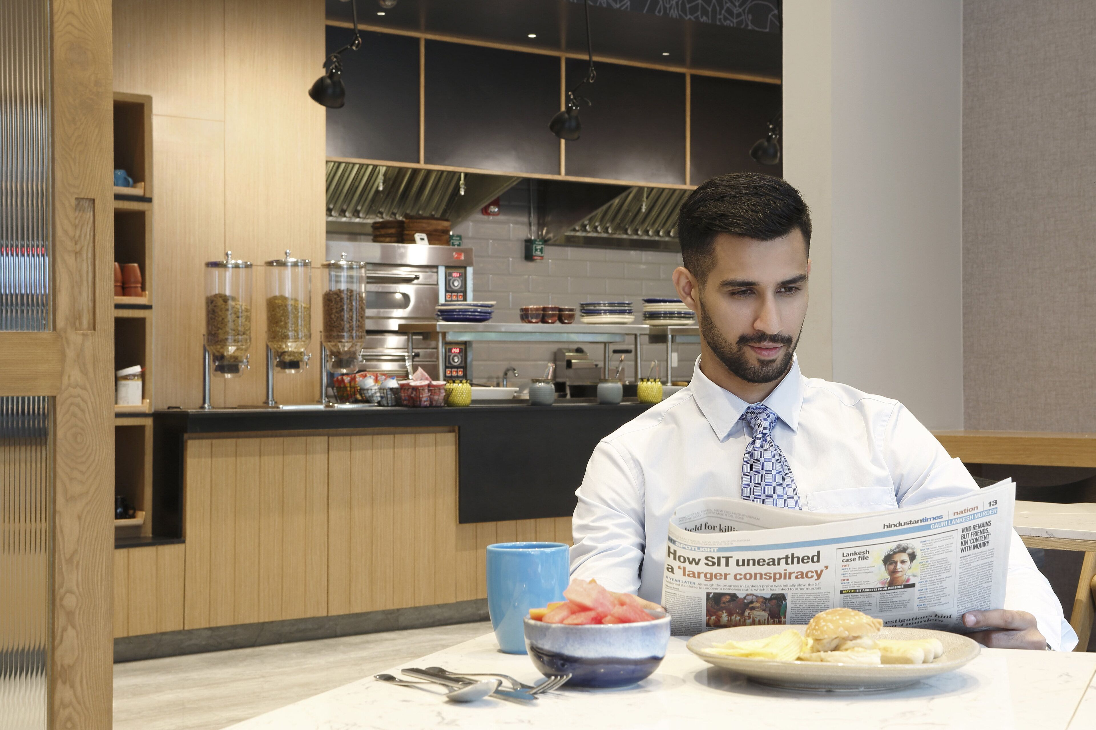 A person reading a newspaper at a table with a bowl of food and a plate of food.