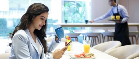 A person sitting at a table with a plate of food, a glass of orange juice, and a blue mug. Another person is in the background near a counter.