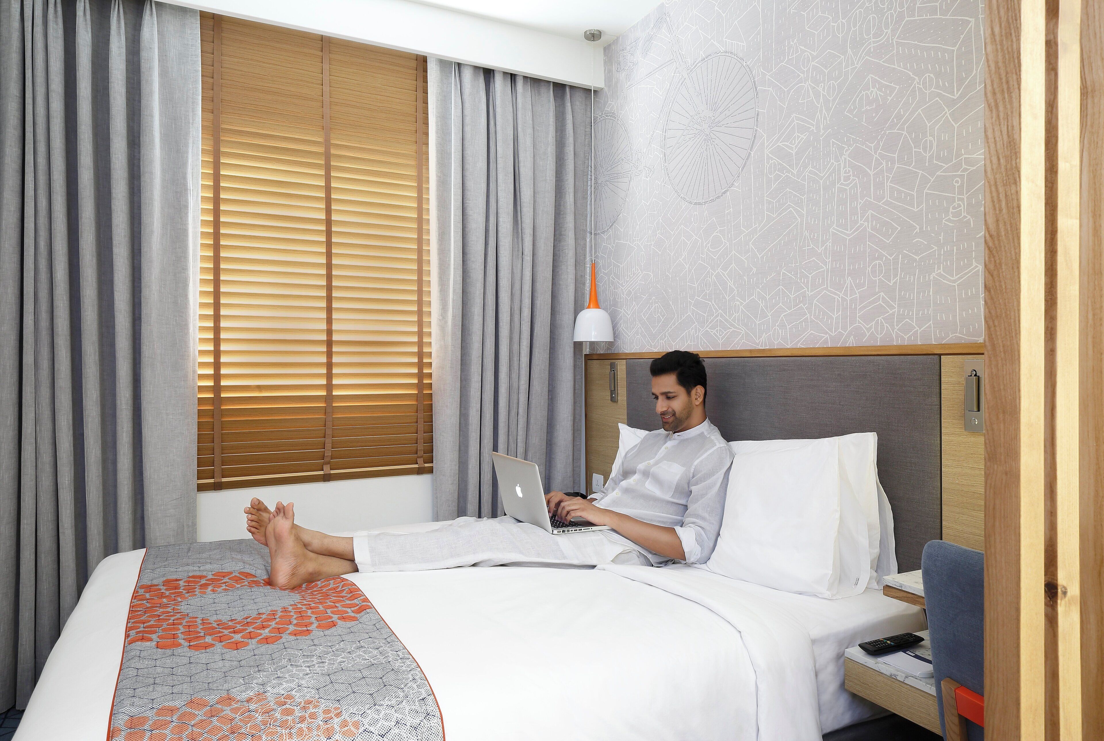 A hotel room with a bed, a laptop, a bedside table, a lamp, and a window with blinds.