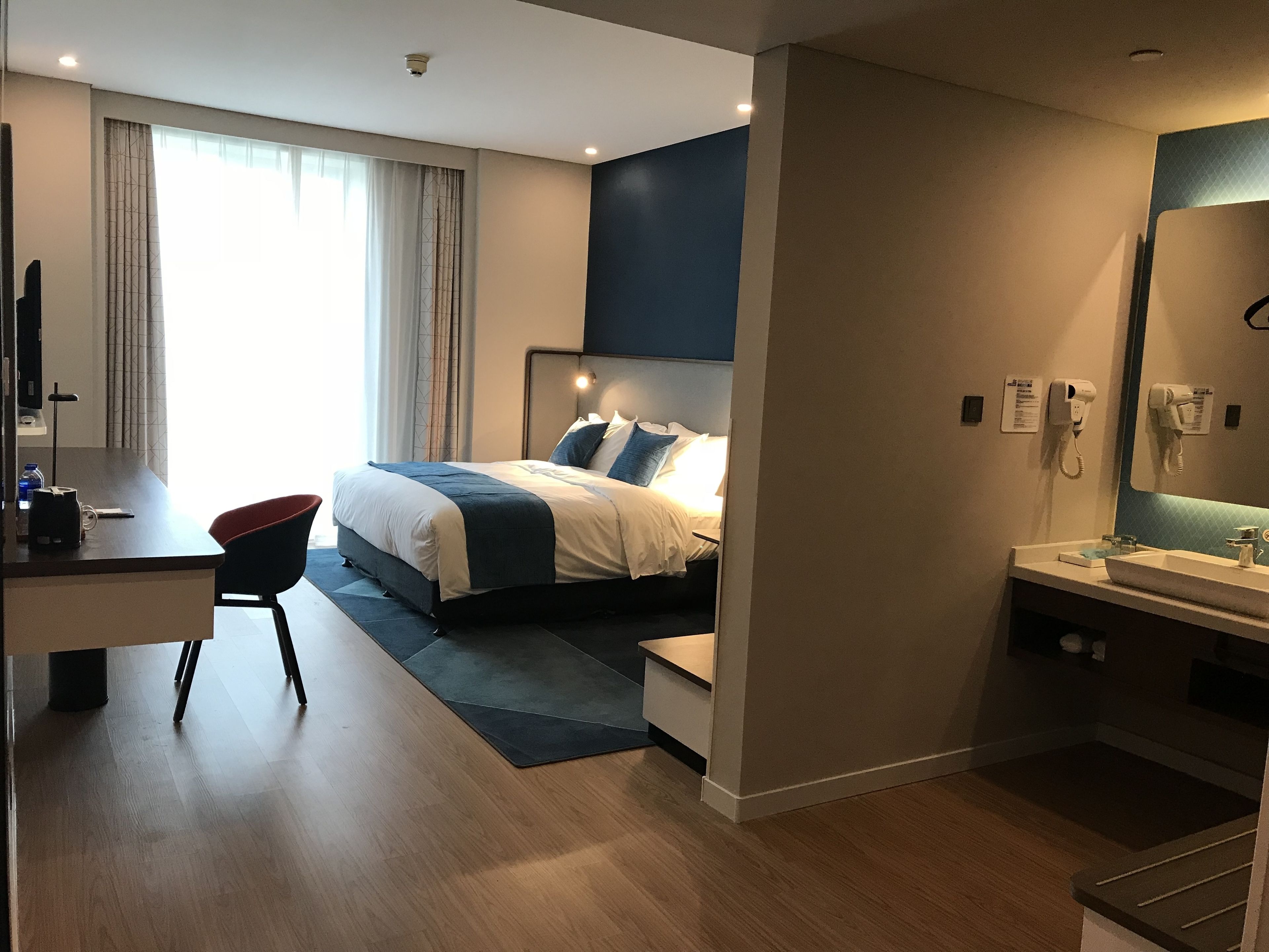 Una habitación de hotel moderna con cama, escritorio, silla y baño con lavabo y espejo.