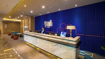Zona de recepción de hotel con mostrador de mármol, una recepcionista y pared azul con diseño geométrico.
