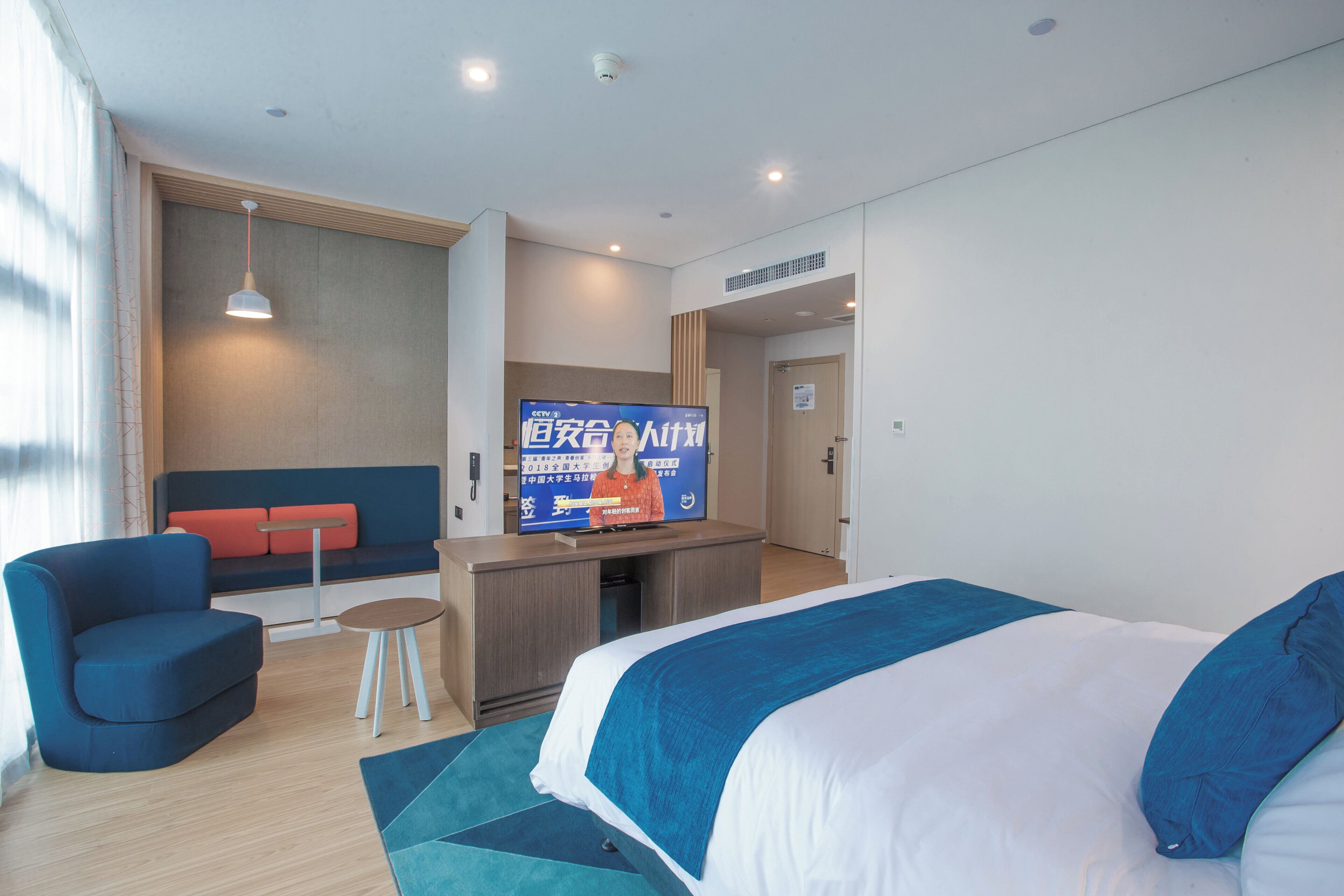 Una camera d'albergo con un letto, una zona salotto con divano e poltrone, un tavolino e una televisione che mostra un canale di notizie.