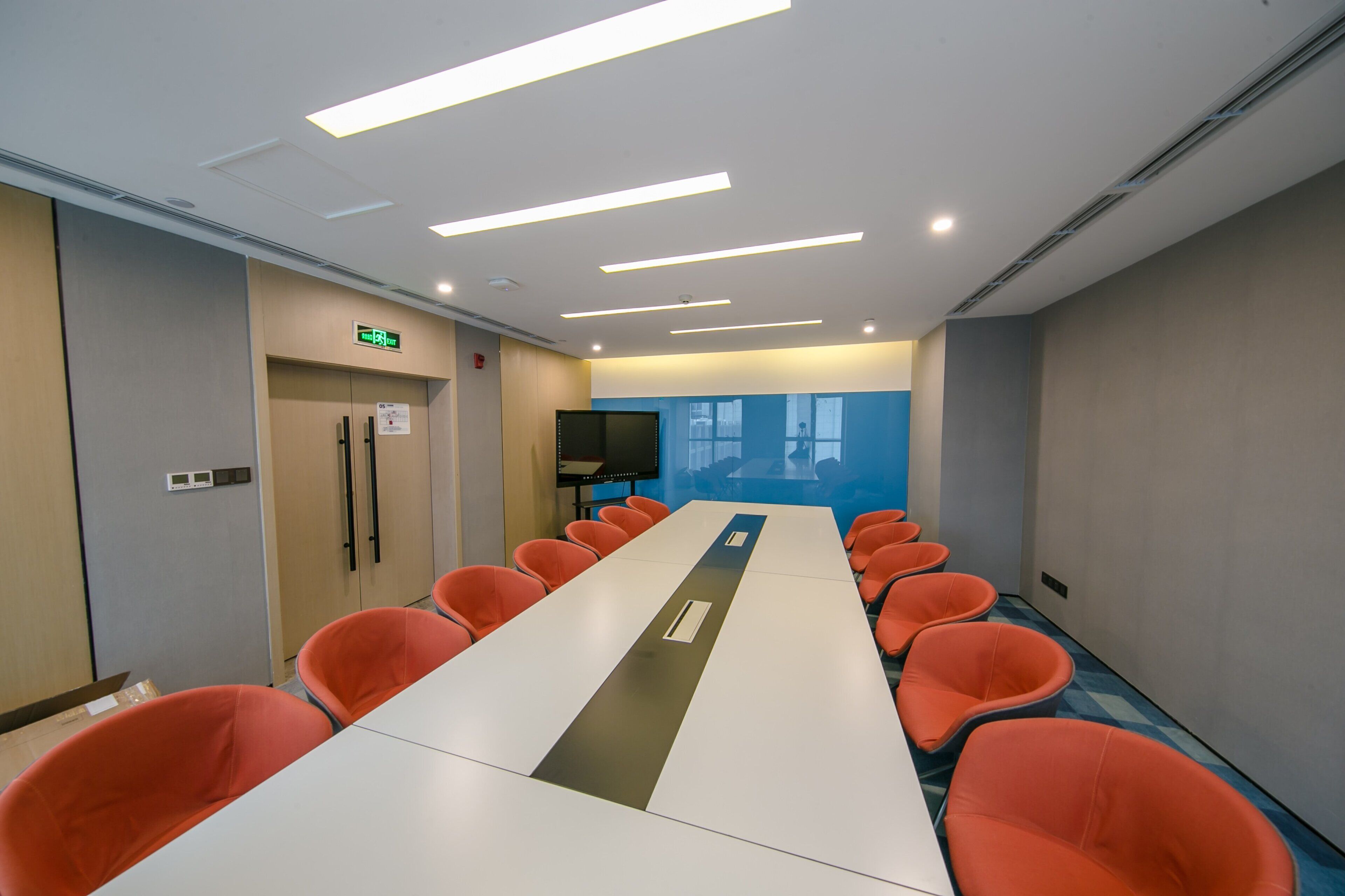 A conference room with a long table, orange chairs, a TV screen, and an exit sign.