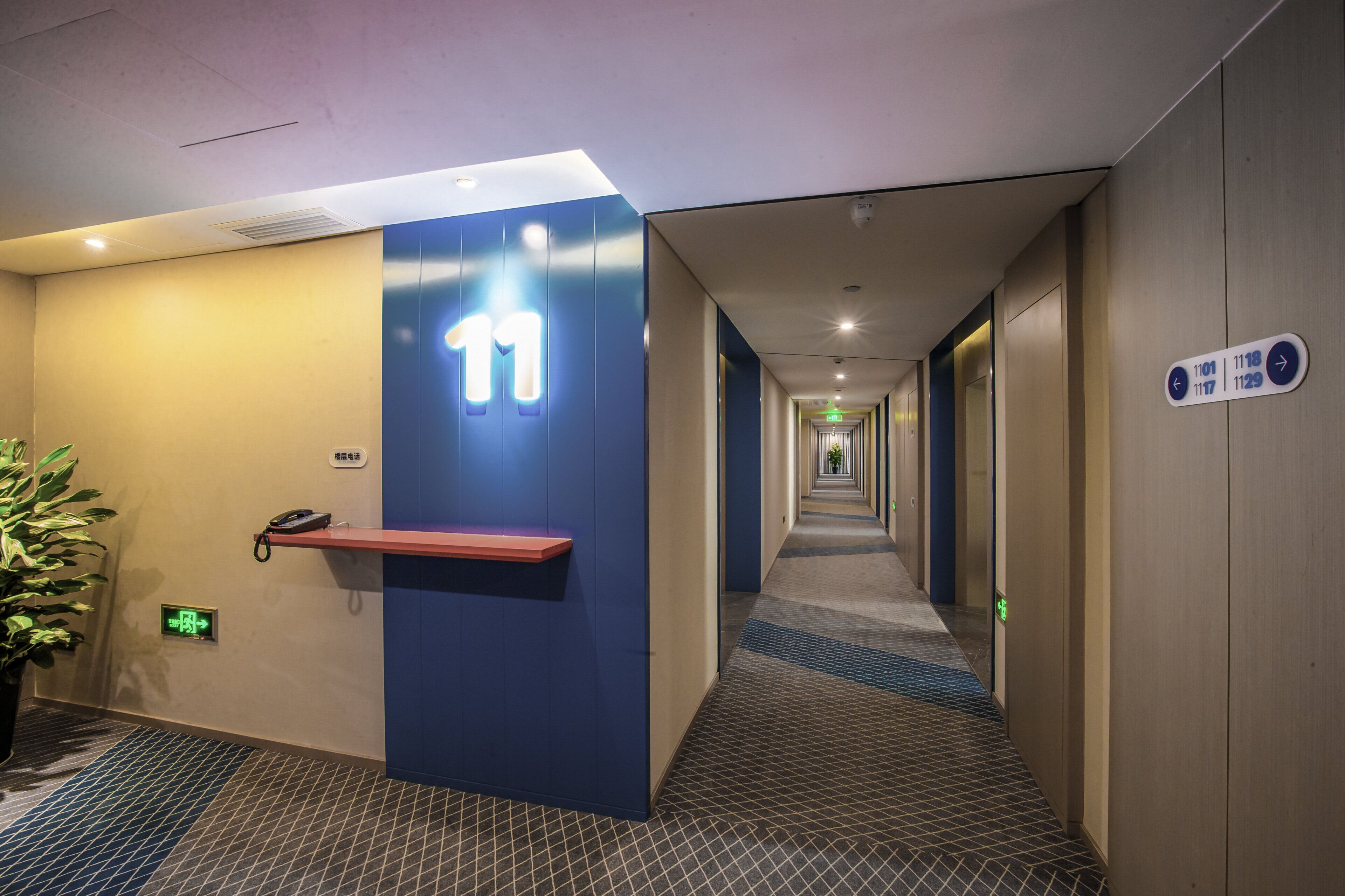 A hotel corridor with a blue wall, a phone on a shelf, and directional signs.
