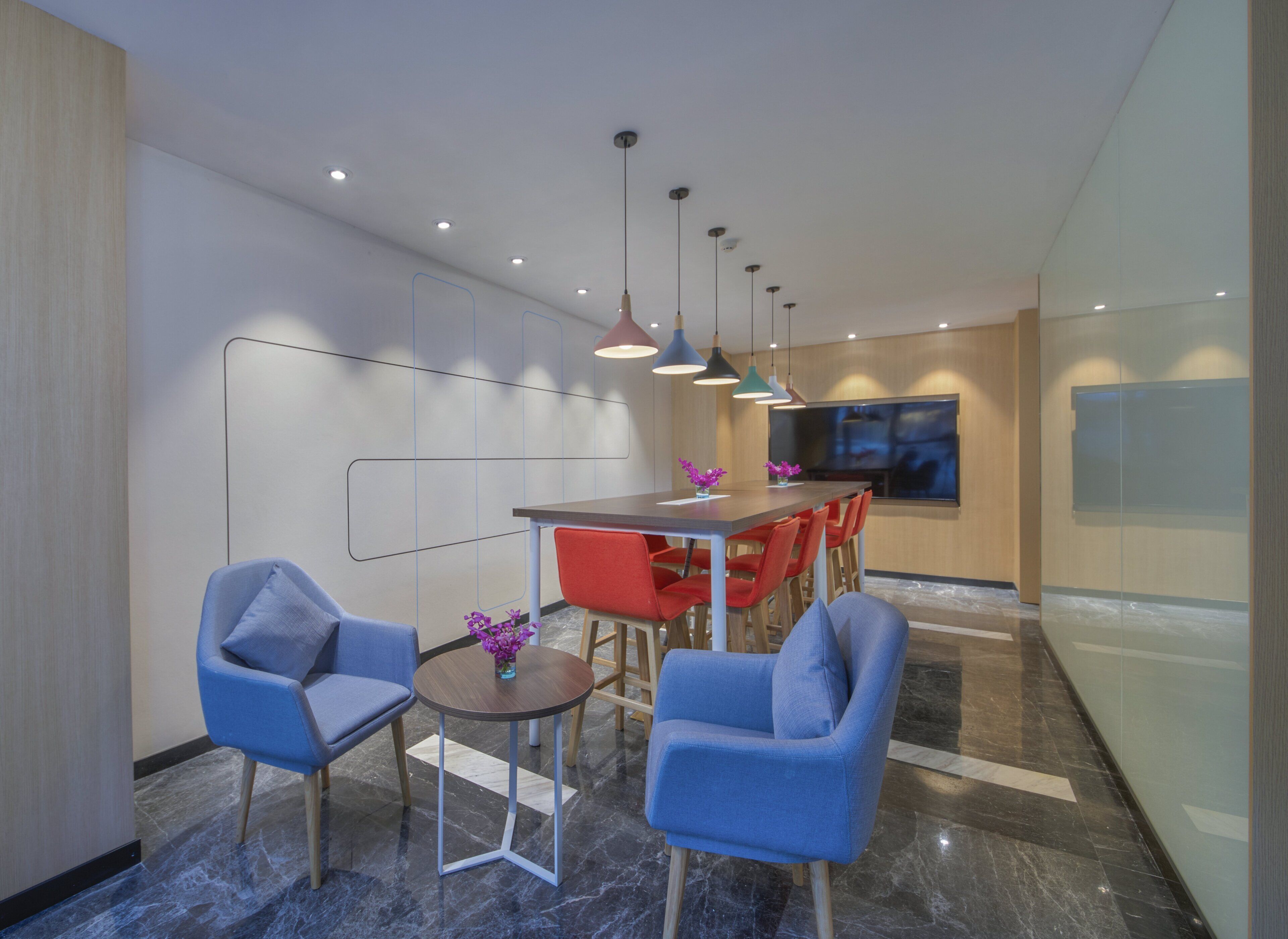 A modern interior with a dining area featuring a wooden table and red chairs, blue armchairs, and pendant lighting.
