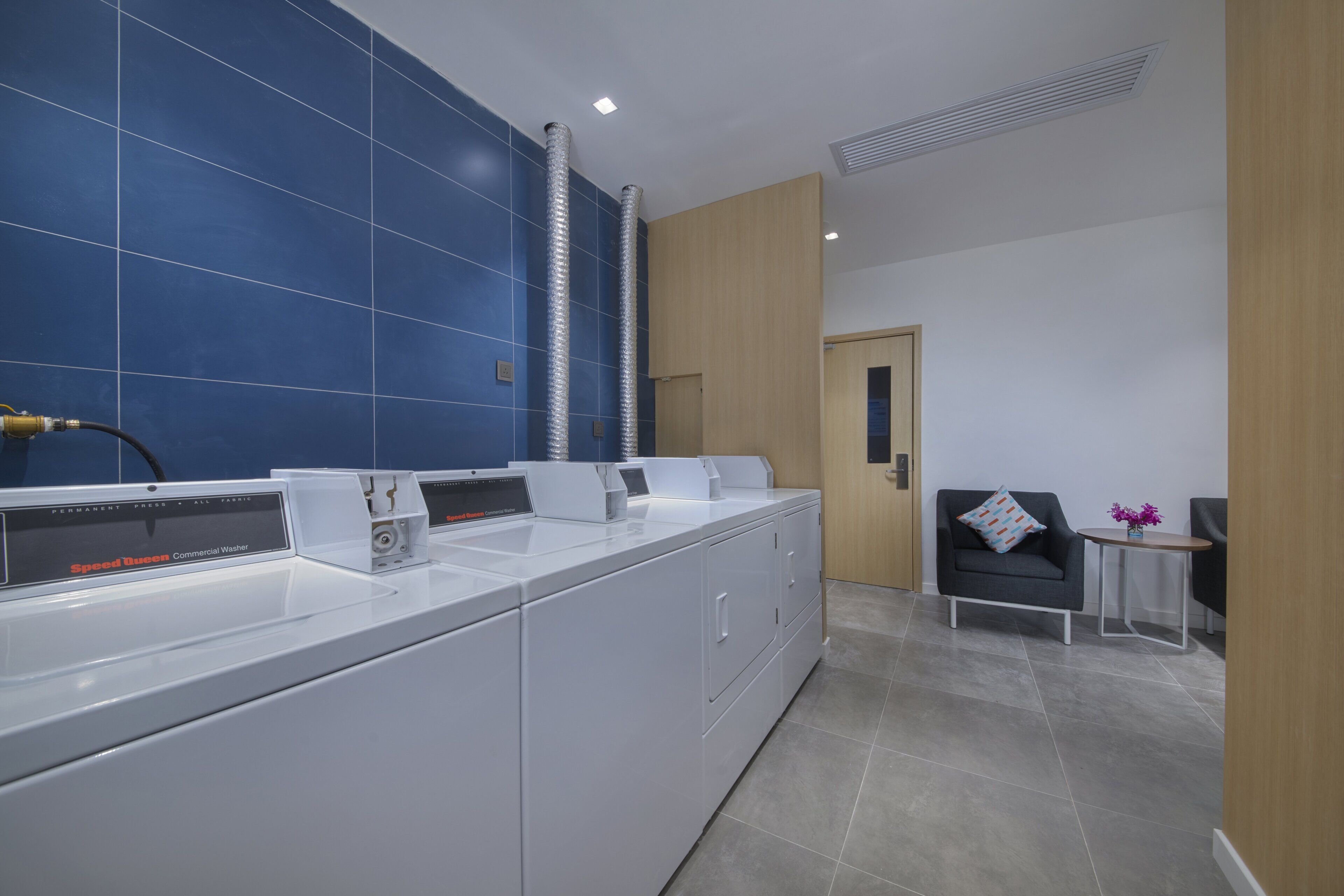 Zona de lavandería comercial con lavadoras, un lavabo y una pequeña área de descanso con mesa y silla.