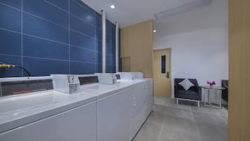 Zona de lavandería comercial con lavadoras, un lavabo y una pequeña área de descanso con mesa y silla.