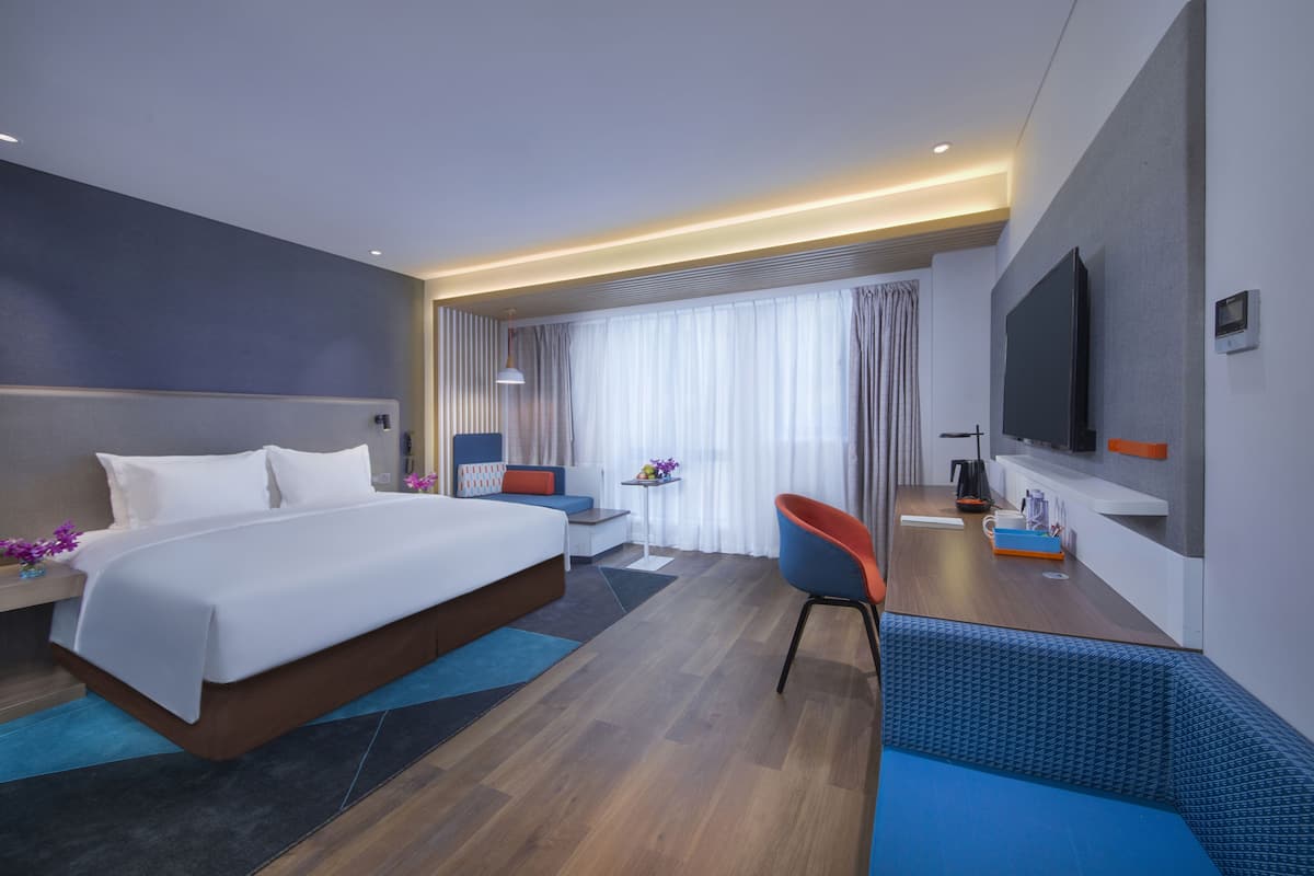 A modern hotel room with a large bed, a desk with a chair, a TV, and a seating area with a sofa.