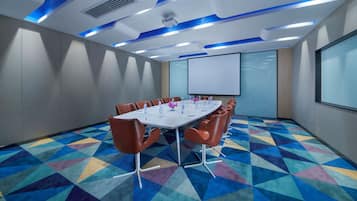 Un salón de conferencias con una mesa blanca, sillas marrones, una pantalla de proyector y una alfombra geométrica de colores.