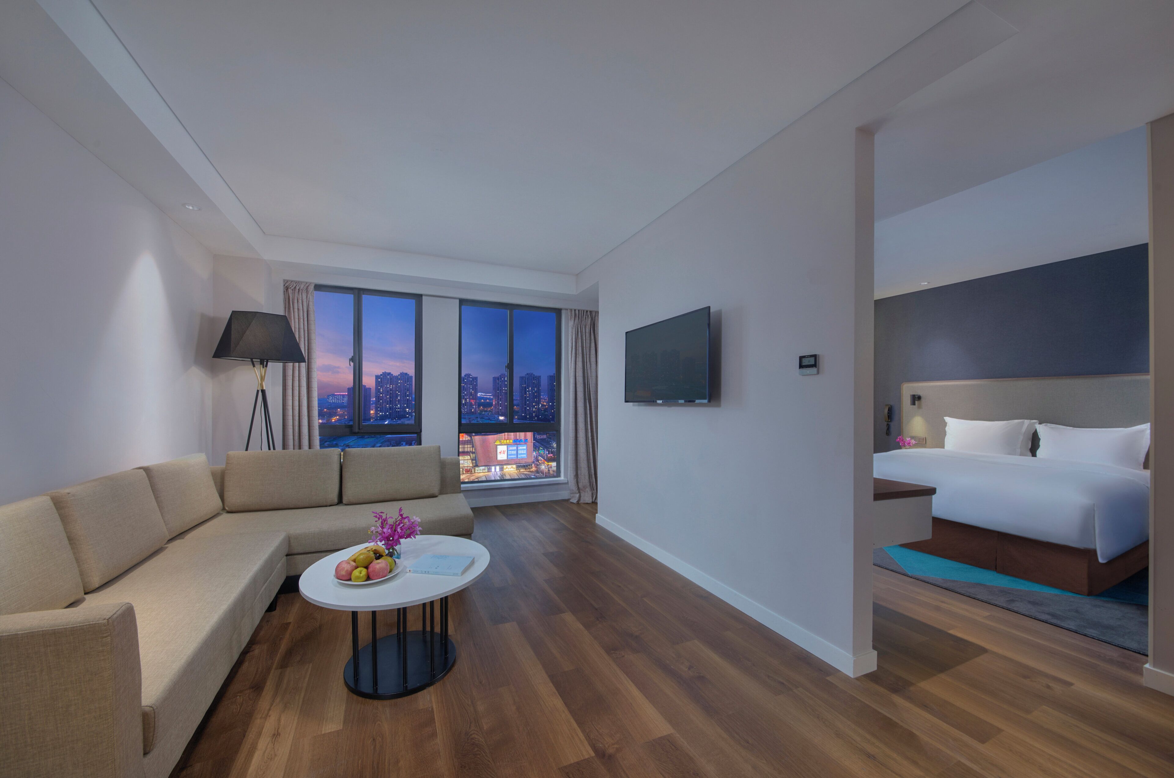 Una habitación de hotel moderna con sofá, una mesa redonda con fruta, cama, televisor y vista a la ciudad a través de ventanas grandes.