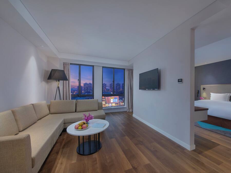 A modern hotel room with a sofa, a round coffee table with fruit, a bed, a TV, and a city view through large windows.
