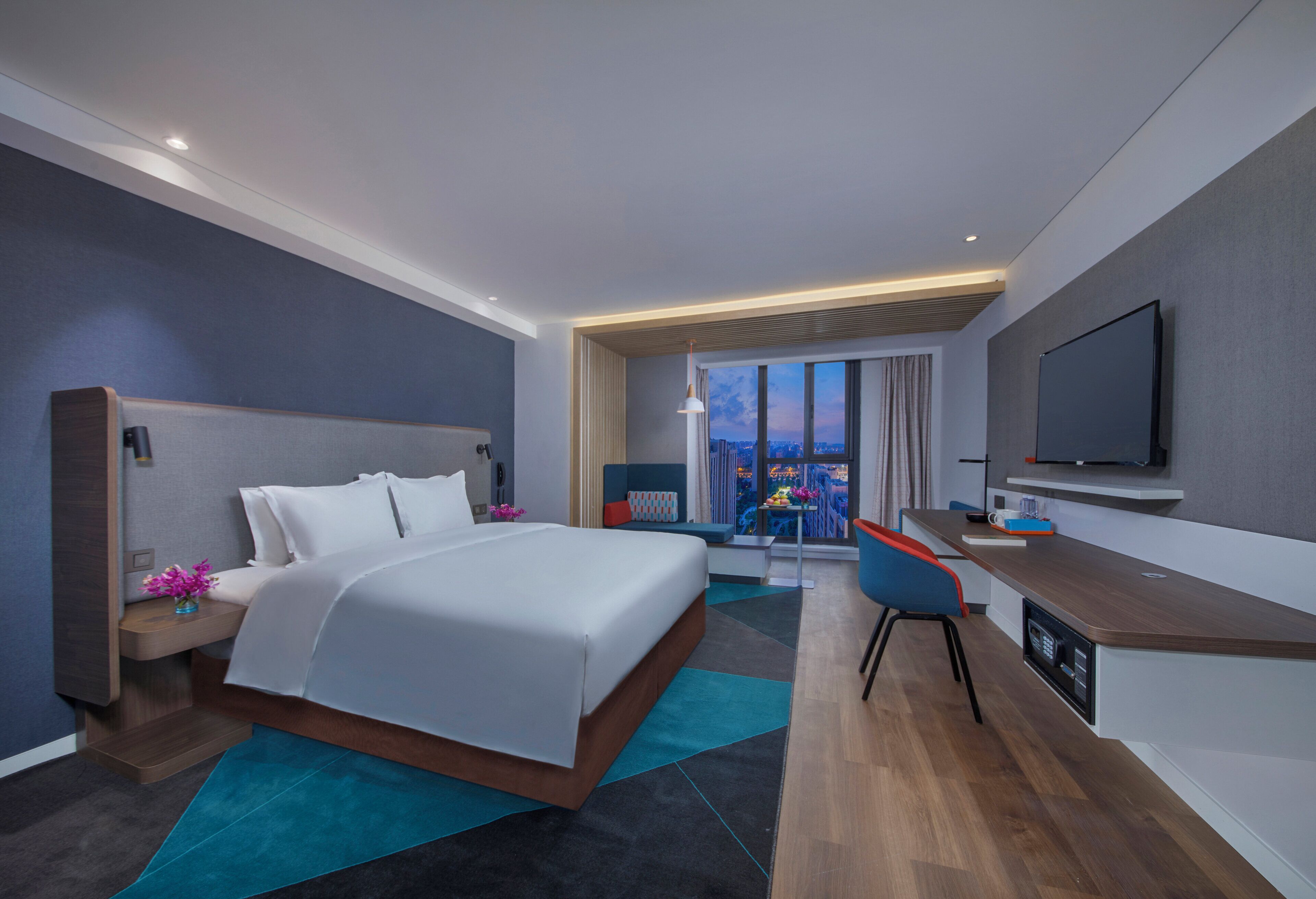 A modern hotel room with a large bed, a desk, a flat-screen TV, and a city view.