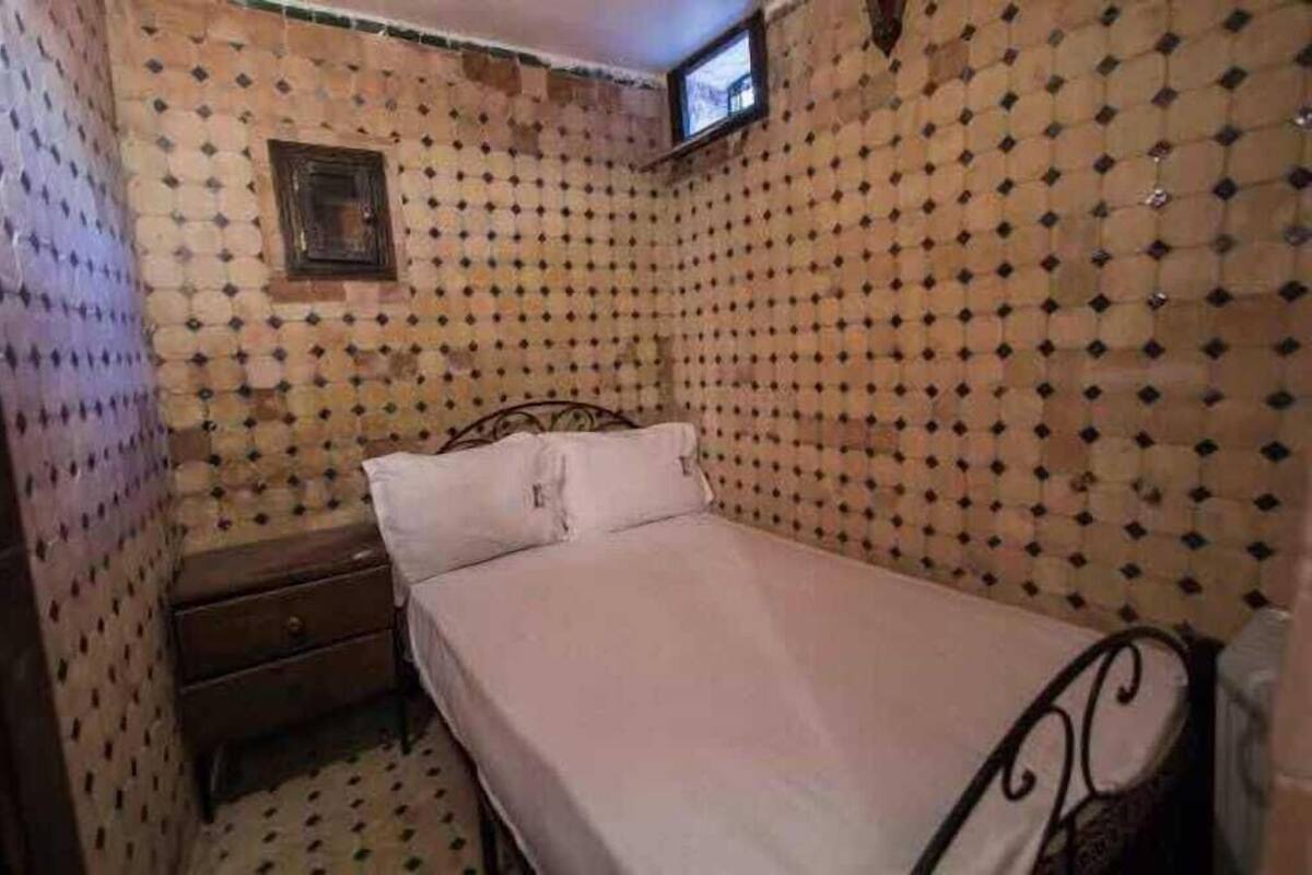 A small, dimly lit room with a single bed, a nightstand, and a small window.