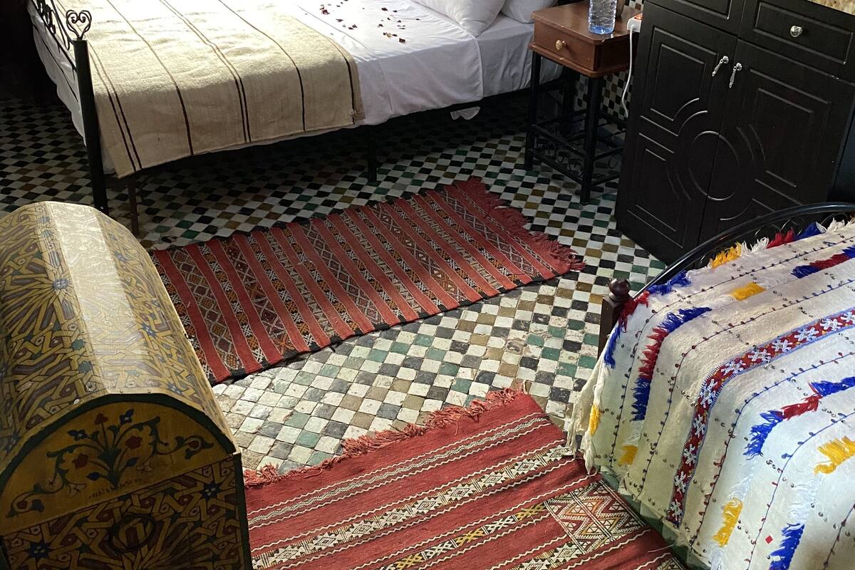 A bedroom with a checkered floor, a bed with white linens, a wooden nightstand, a dresser, and a patterned rug.