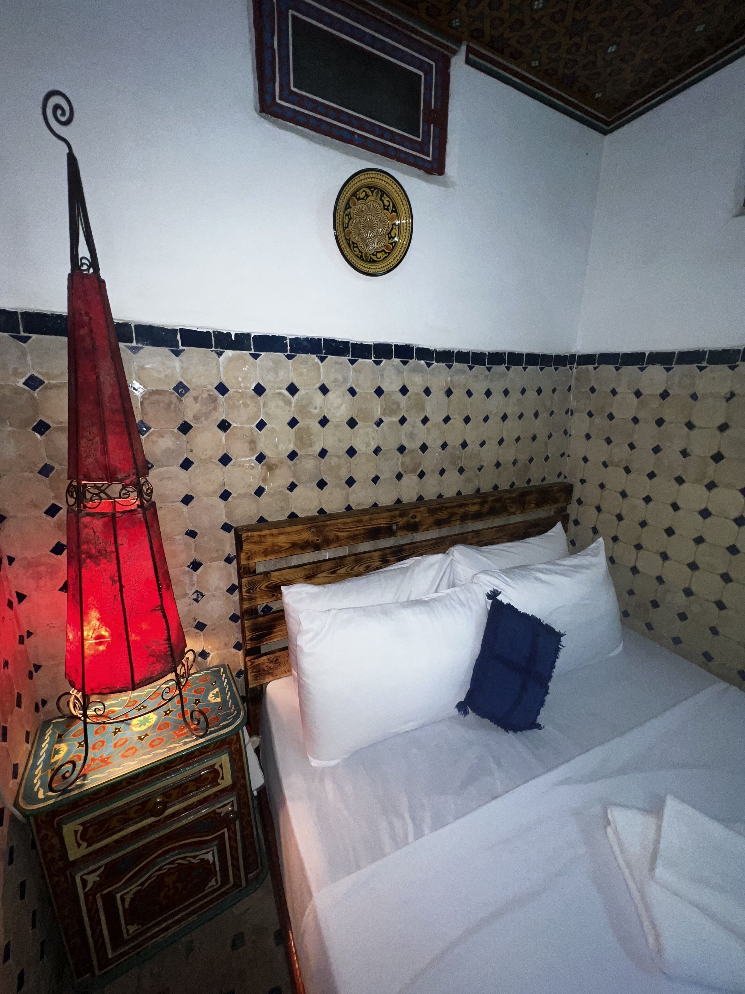 A bed with white and blue pillows, a red hanging lamp, and a decorative plate on the wall.