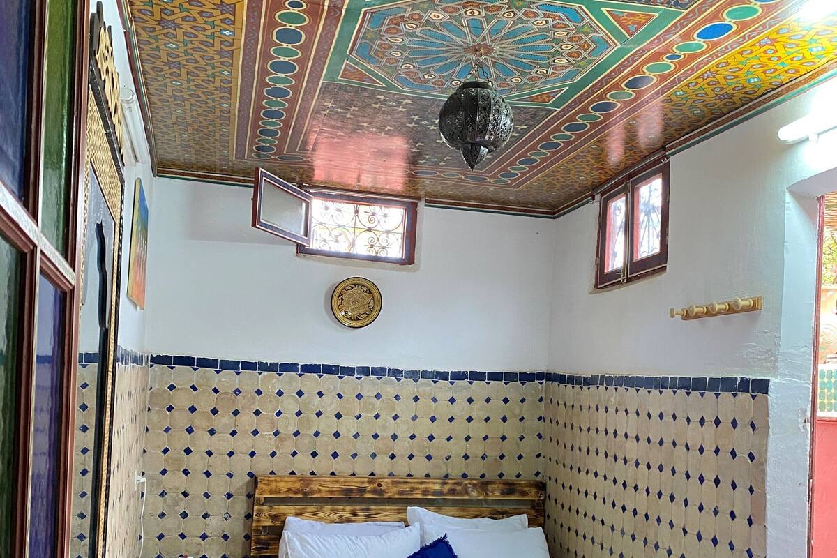 A room with a patterned ceiling, a bed with white bedding, a wooden headboard, a small bedside table, and a wall-mounted light fixture.
