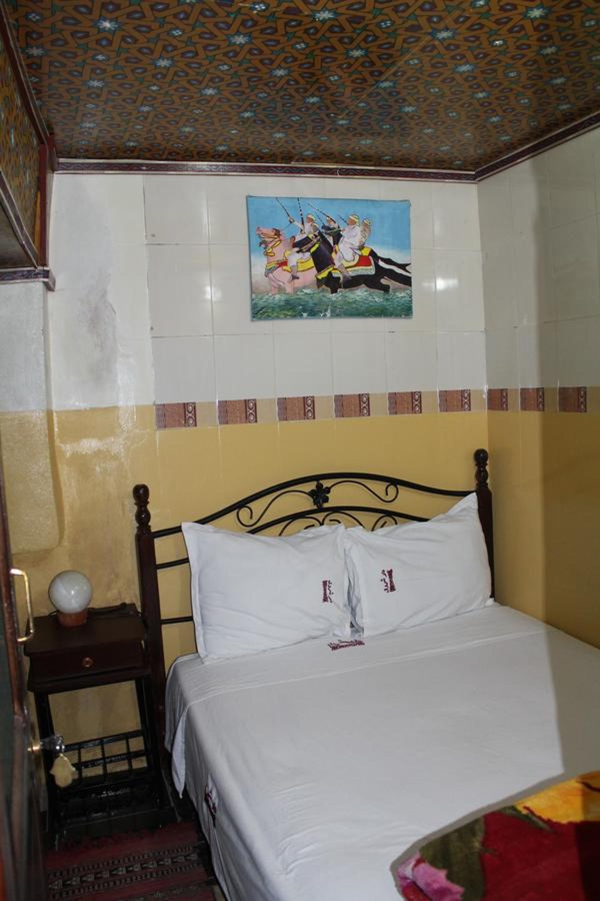 Een slaapkamer met een bed, een nachtkastje, een aan de muur hangend schilderij en een kamer met een patroonplafond.