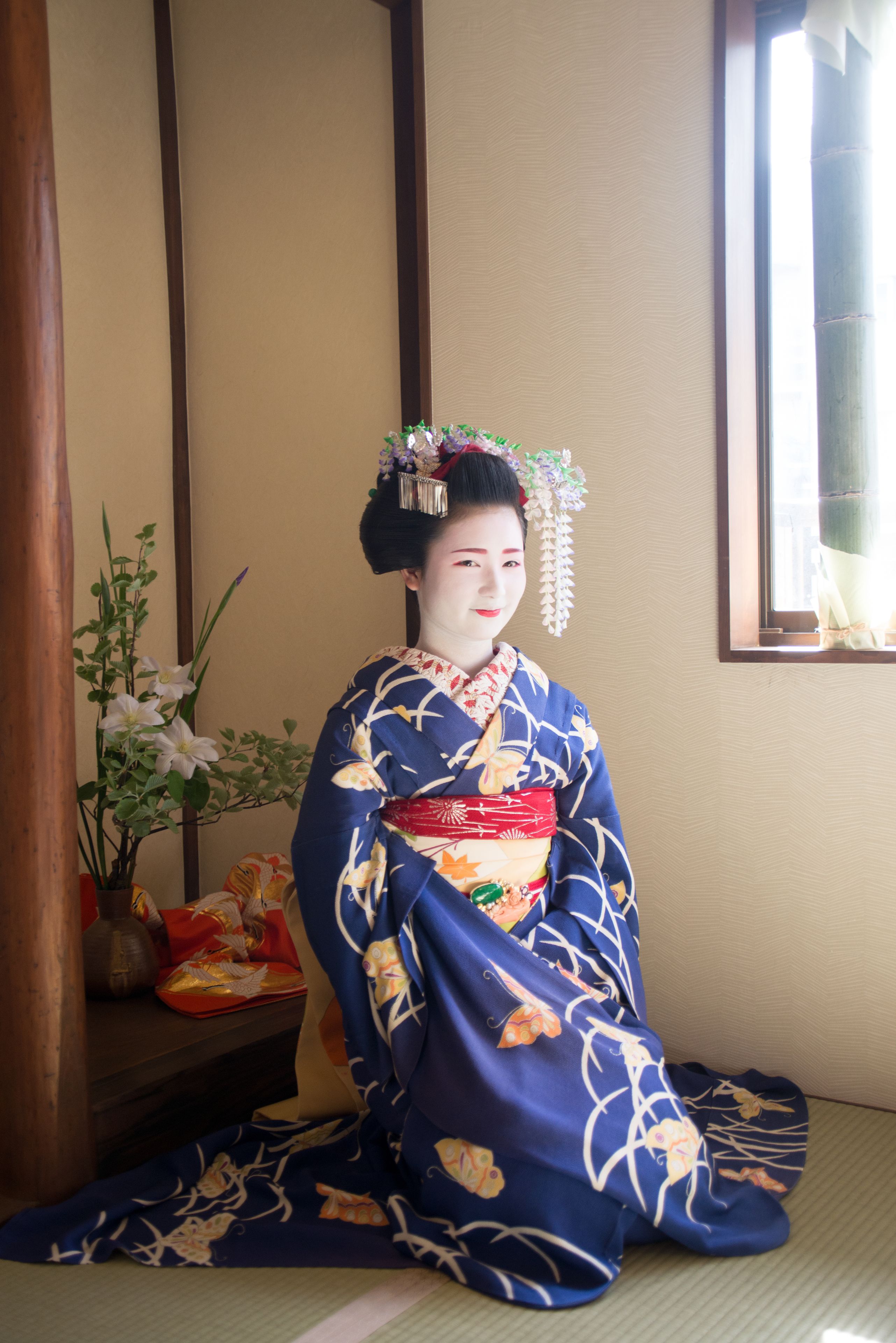 A traditional Japanese room with tatami flooring, a low wooden table with a vase of flowers, and a window with a view of the outside.