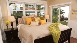 A bedroom with a bed, two nightstands, a window with a view of palm trees, and a door to a balcony.