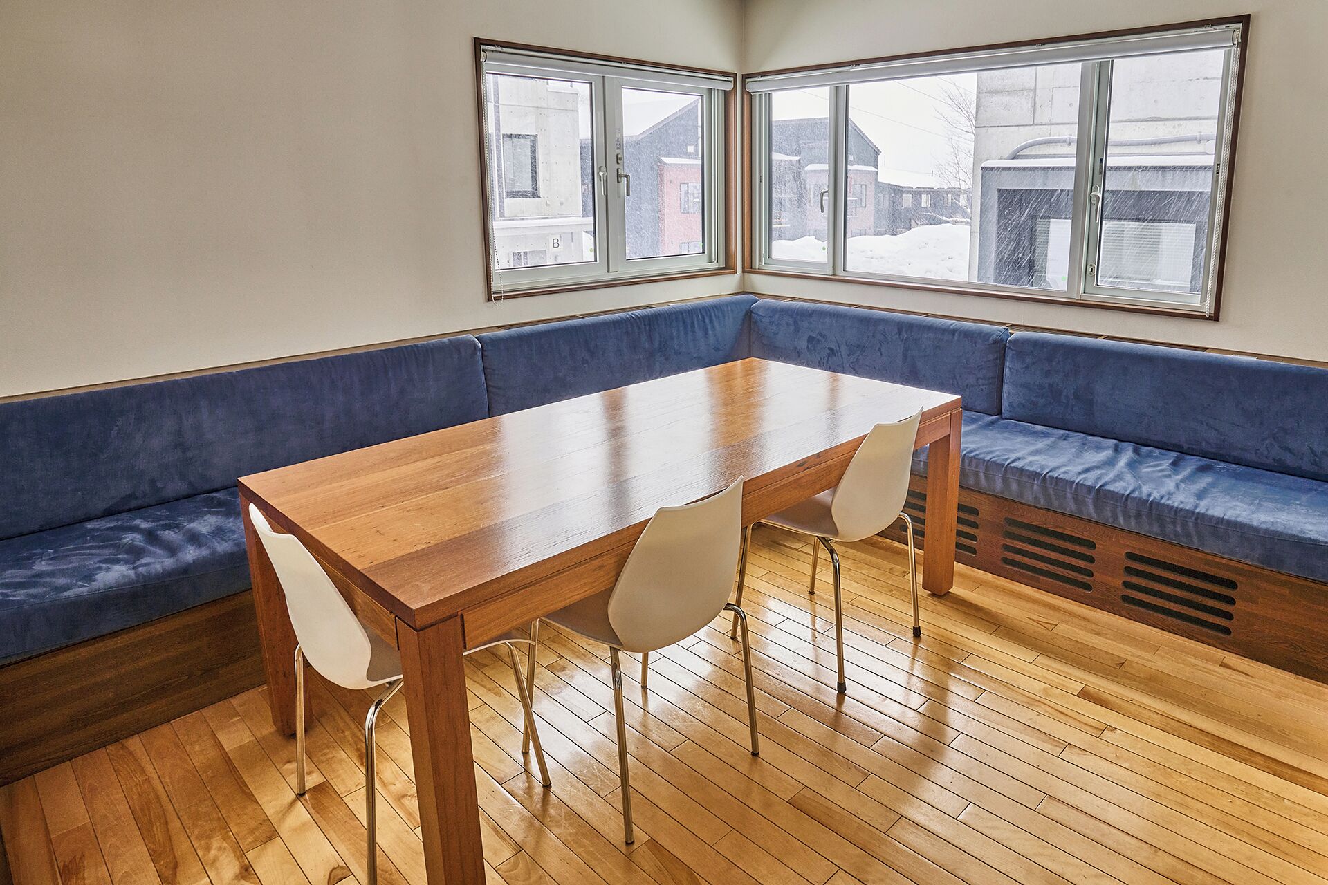 A corner room with a wooden table and chairs, a blue cushioned bench, and large windows with a view of buildings outside.