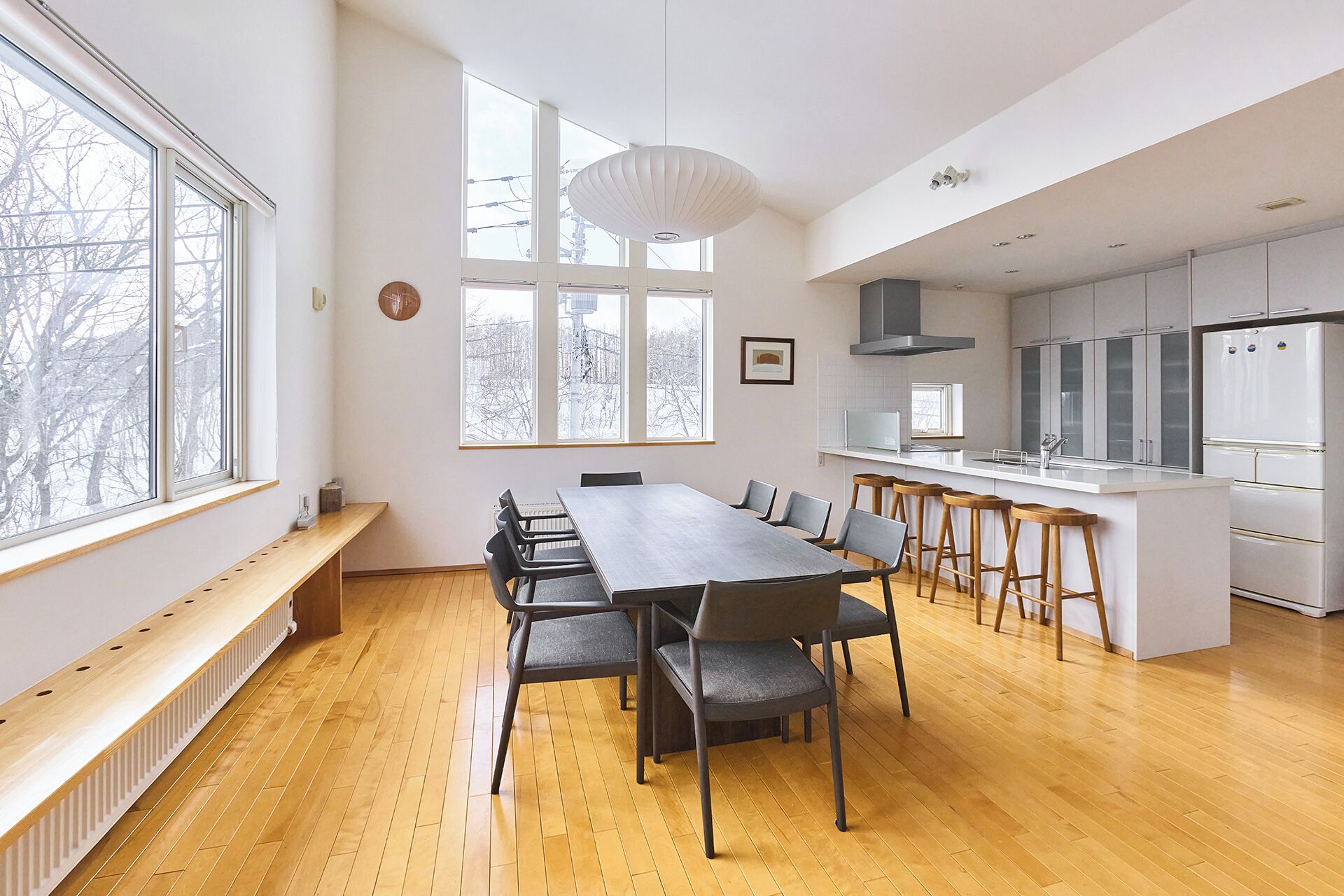 Una cocina moderna con una isla central, una zona de comedor con una mesa larga y ventanas amplias que permiten la entrada de luz natural.