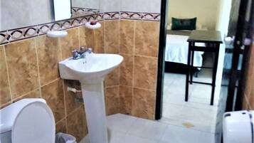 Un baño con inodoro, lavabo y espejo. En la habitación contigua hay una cama y una mesa.