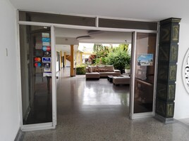 Un vestíbulo de hotel con puerta de vidrio de entrada, zona de estar con sofá y reposapiés, y un cartel en la pared.