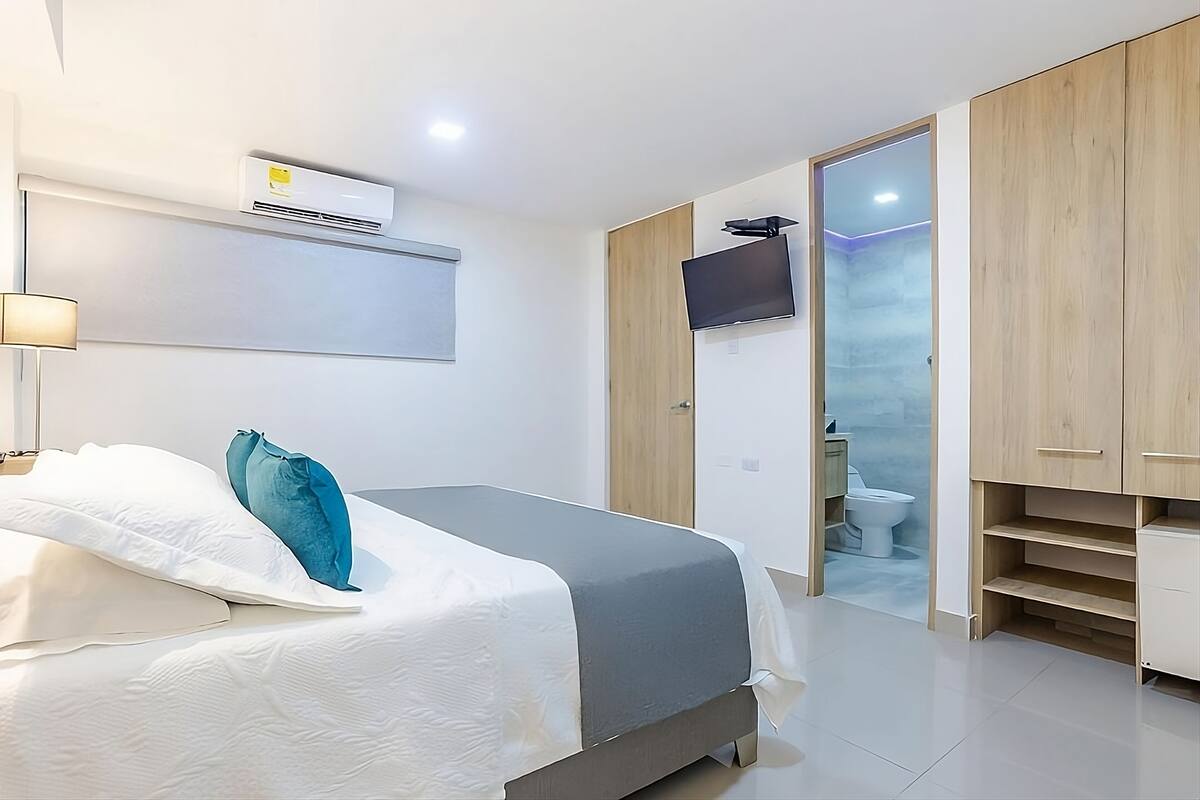 Une chambre moderne équipée d’un lit, d’un téléviseur à écran plat, d’un appareil de climatisation et d’une salle de bain visible à travers une porte ouverte.