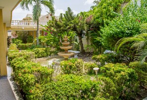 Un jardín bien cuidado con una fuente escalonada, abundante vegetación y un balcón con plantas en macetas.