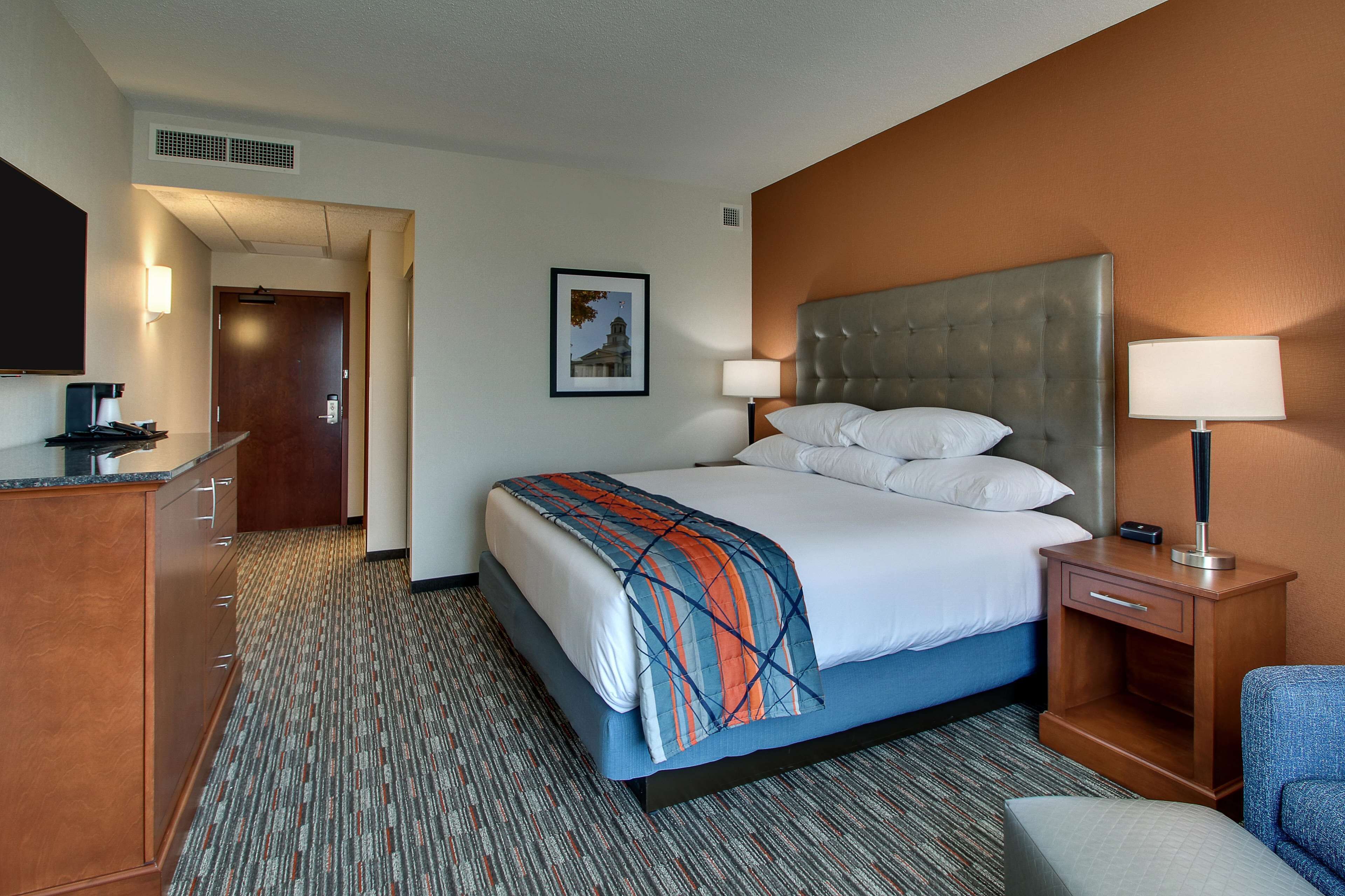 A hotel room with a large bed, a desk with a chair, a nightstand with a lamp, and a picture on the wall.
