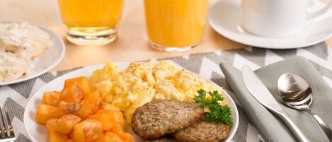 Un plato de desayuno con huevos revueltos, croquetas de patata y salchichas, acompañado de un vaso de zumo de naranja y una taza de café.